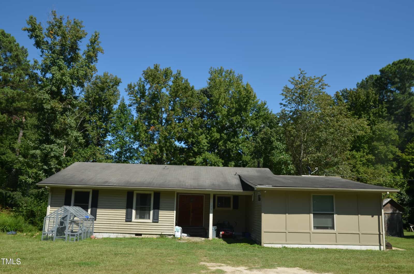 A single-story house with beige siding and a dark roof sits in front of a dense group of trees. There is a small greenhouse on the grass to the left and some items near the front door. The sky is clear and blue.