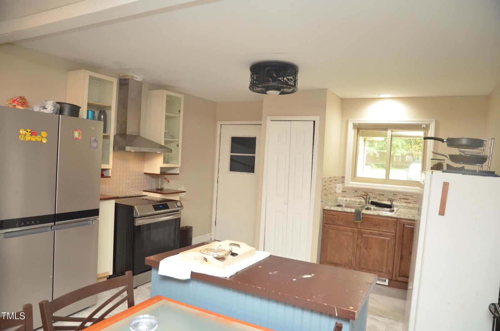 A kitchen with brown cabinets, stainless steel appliances, a fridge with magnets, a small island with papers on top, a window above the sink, and a door with a frosted window.