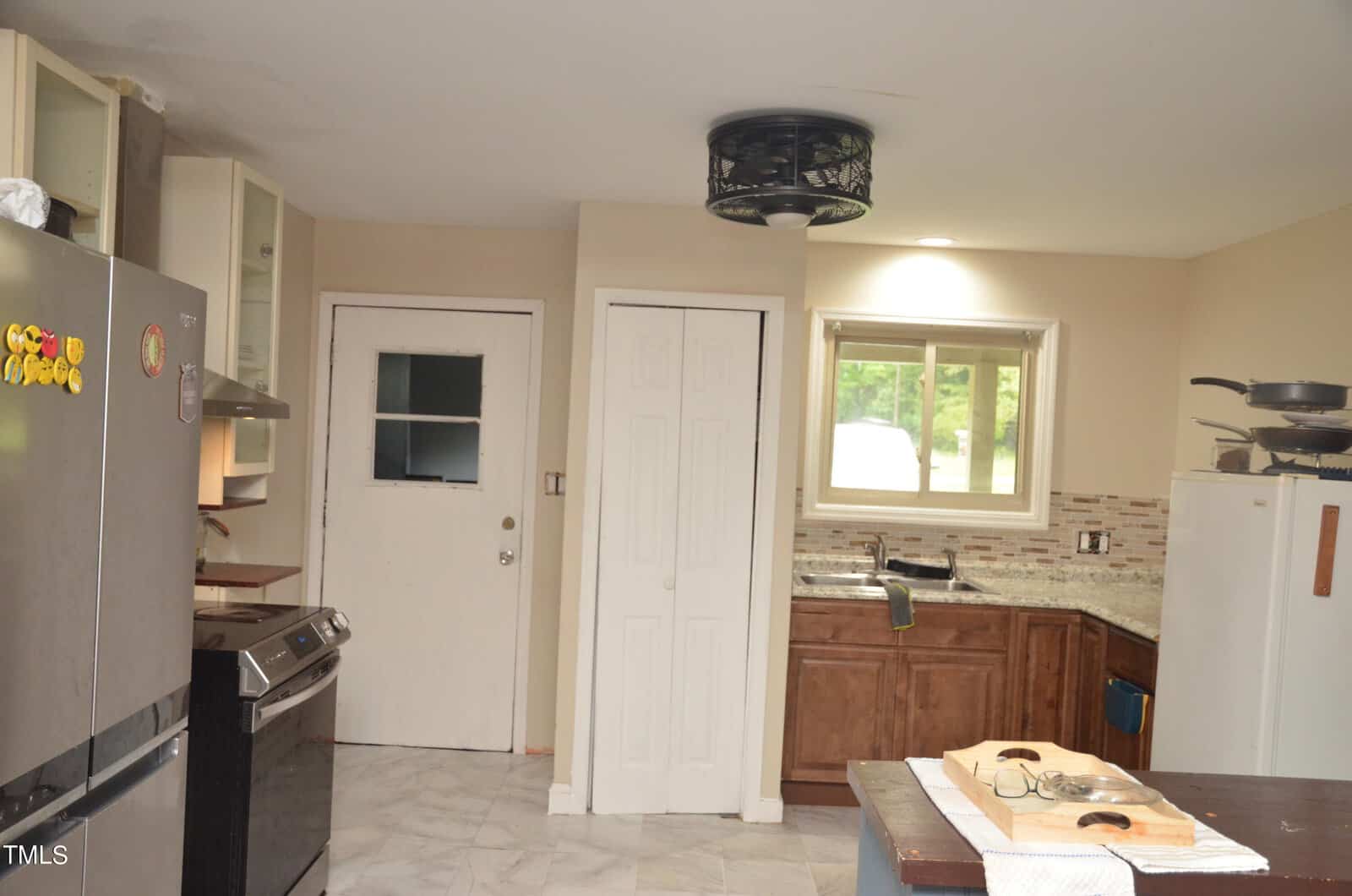 A kitchen with light-colored tile floors, beige walls, dark wood cabinets, and granite countertops. There is a stove, refrigerator, sink under a window, shelves, and a white door with a window. A table is partially visible.