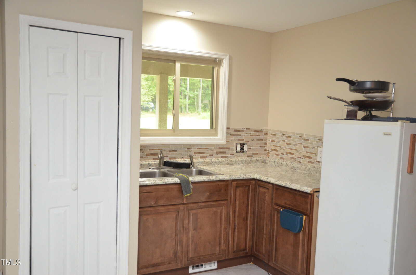 A small kitchen with brown cabinets, a beige tiled backsplash, a window over the sink, a white refrigerator with pans stacked on top, and a white closet door on the left. A towel hangs over the sink.