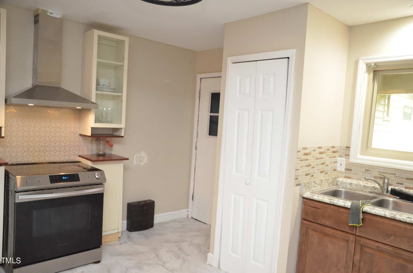 A kitchen with a stainless steel stove, range hood, beige walls, white tile floor, brown cabinets, double sink, tiled backsplash, small window, and a white closet door. Shelves and a small trash bin are also visible.