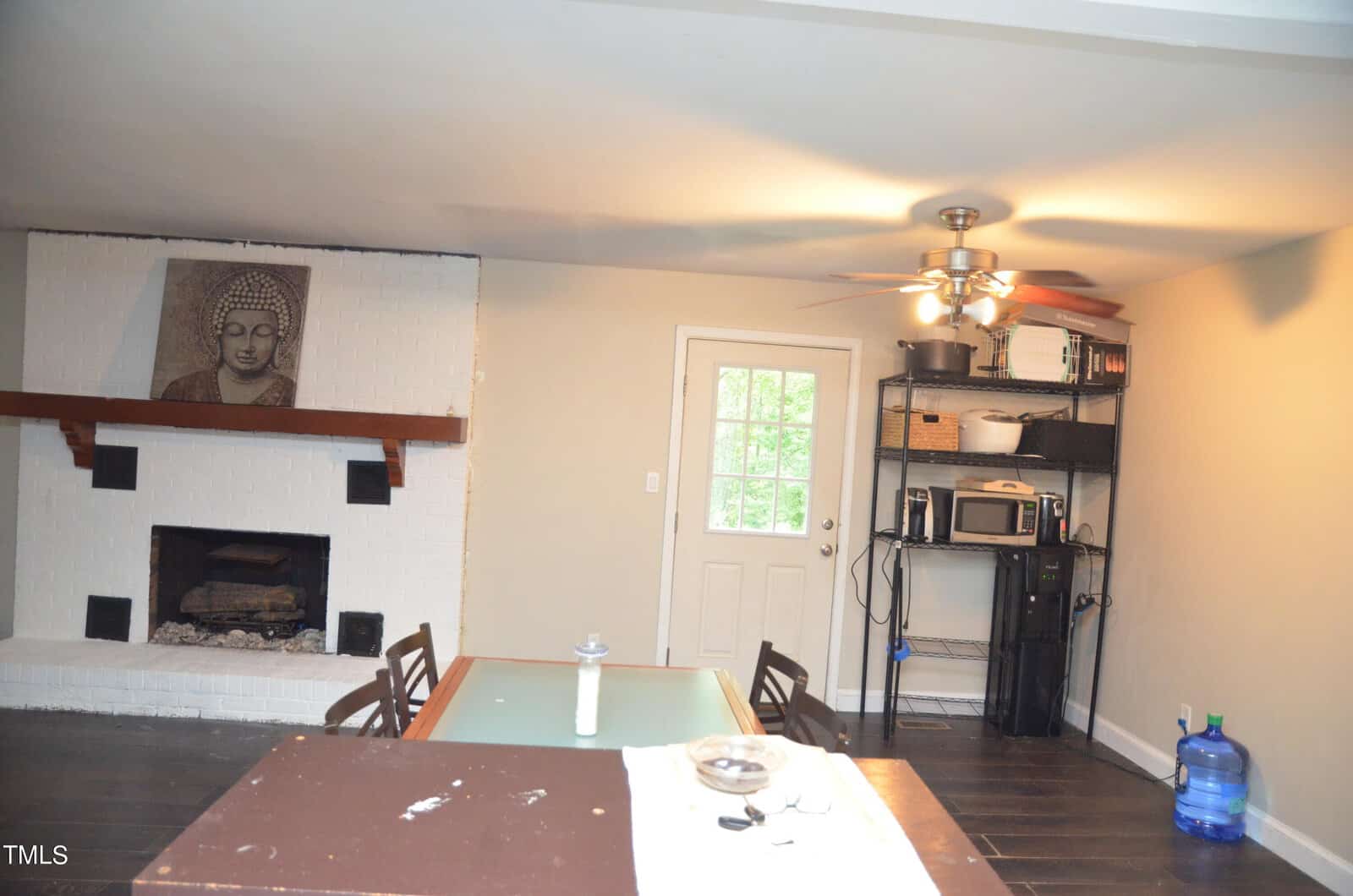 A dining area with a table and chairs, a white brick fireplace featuring a Buddha artwork, a metal shelf holding kitchen appliances, a water jug, and a ceiling fan with lights.