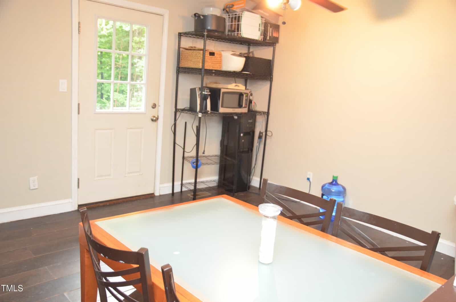 A small dining area with a glass-top table and four chairs, a shelving unit holding kitchen appliances, a water jug, and a door with a window leading outside.