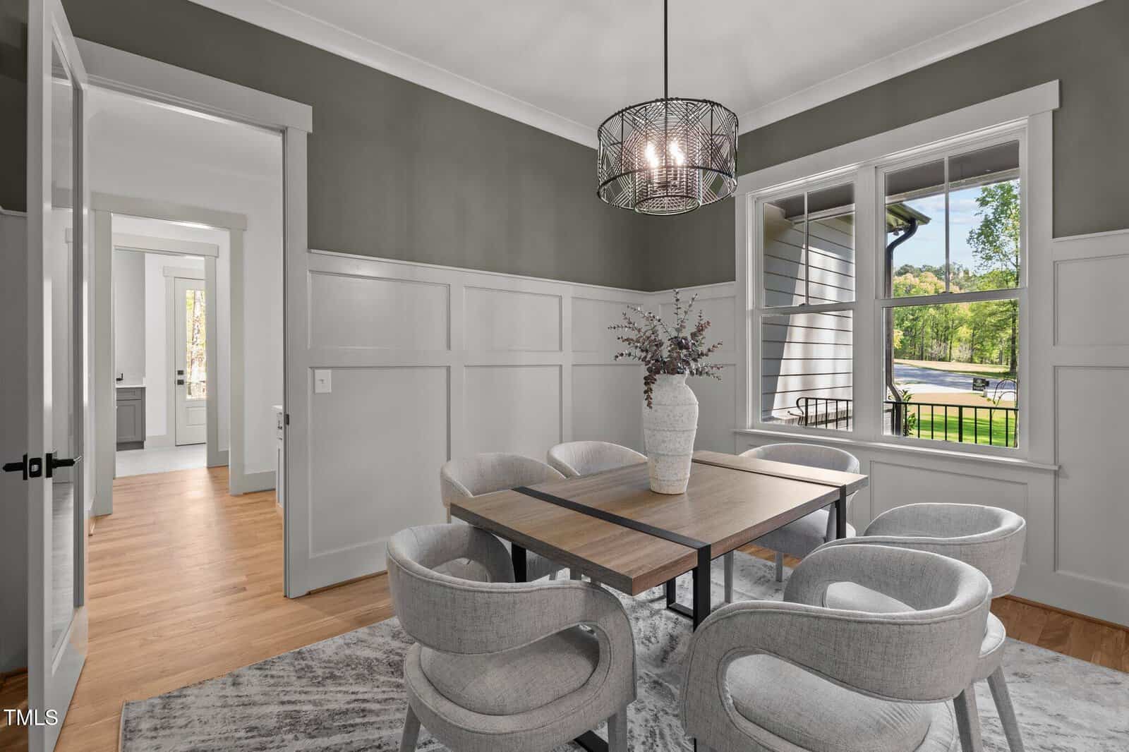 A modern dining room with a wooden table, six upholstered chairs, a large rug, a decorative vase with branches, a black pendant light, green and white paneled walls, wood flooring, and large windows with an outdoor view.