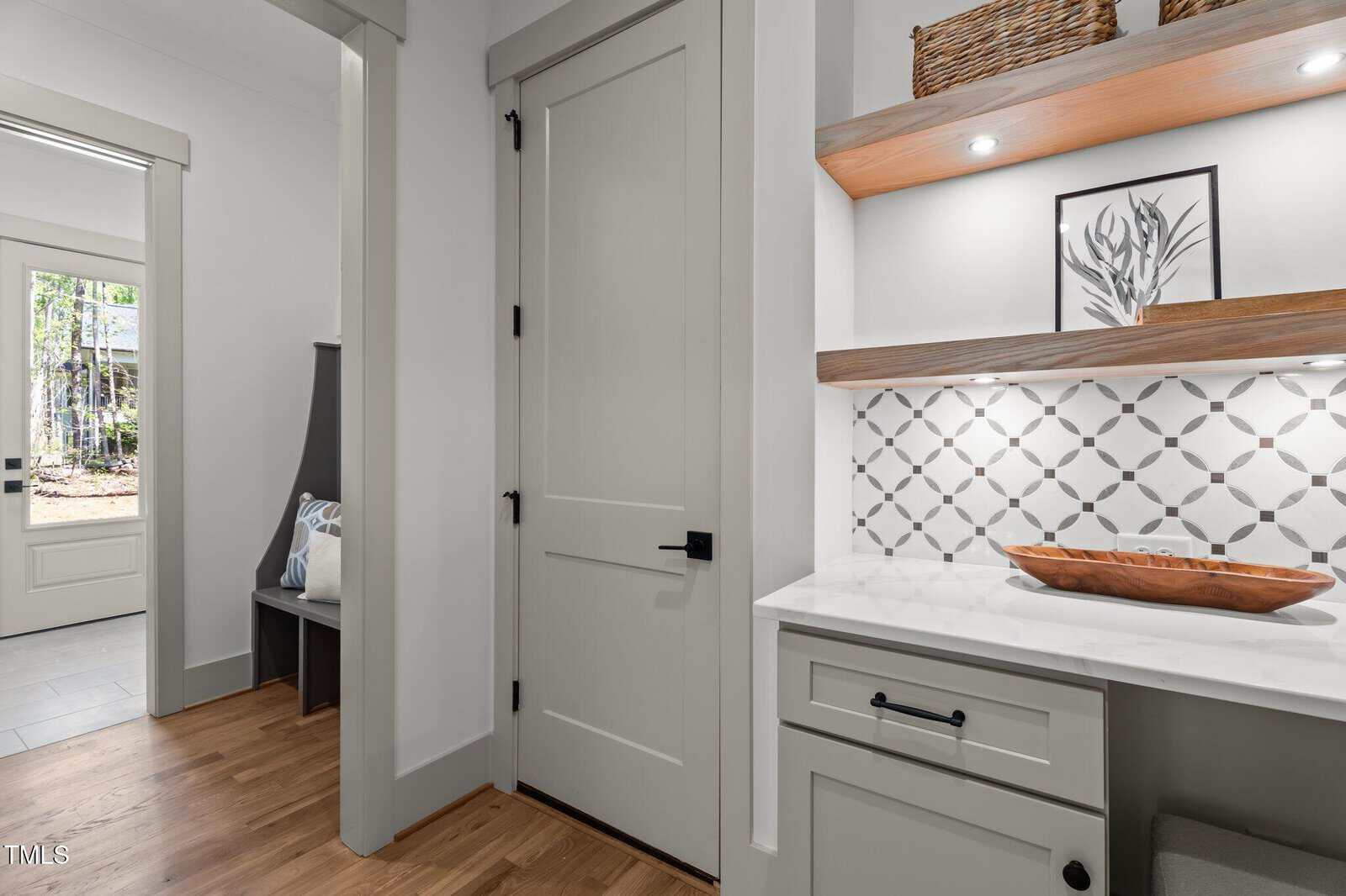 A small modern nook with white cabinets, geometric backsplash, wooden tray, framed art, shelves, and wicker basket. A hallway with a bench and pillows leads to a door with windows showing trees outside.