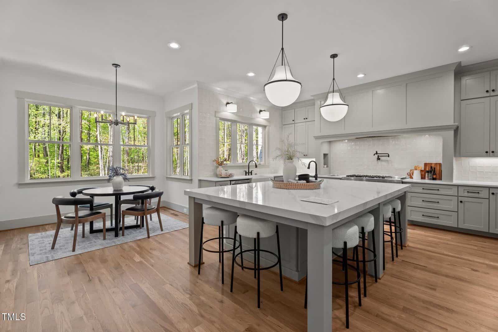 A modern kitchen with a large white island, gray cabinets, barstools, pendant lights, hardwood floors, and a round dining table by large windows overlooking a wooded area.