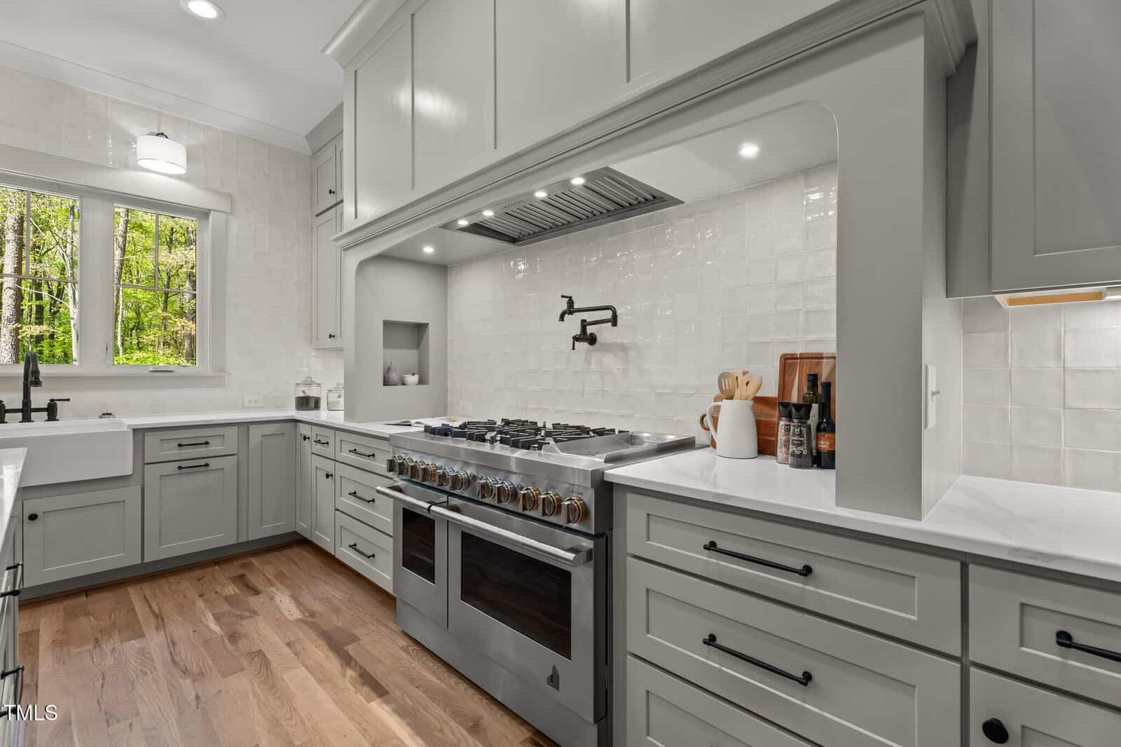 Modern kitchen with light gray cabinets, white countertops, a large stainless steel stove with double ovens, a pot filler faucet, white farmhouse sink under a window, and light wood flooring.