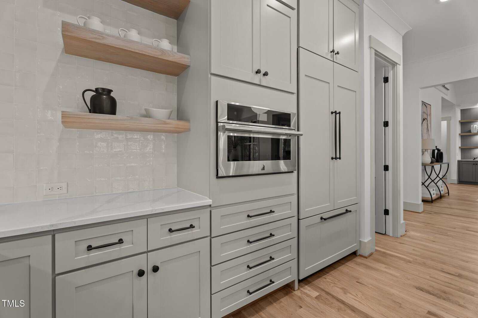 Modern kitchen with white cabinetry, a built-in stainless steel oven, open wooden shelves holding mugs and a teapot, light wood flooring, and a white tile backsplash. A hallway is visible in the background.