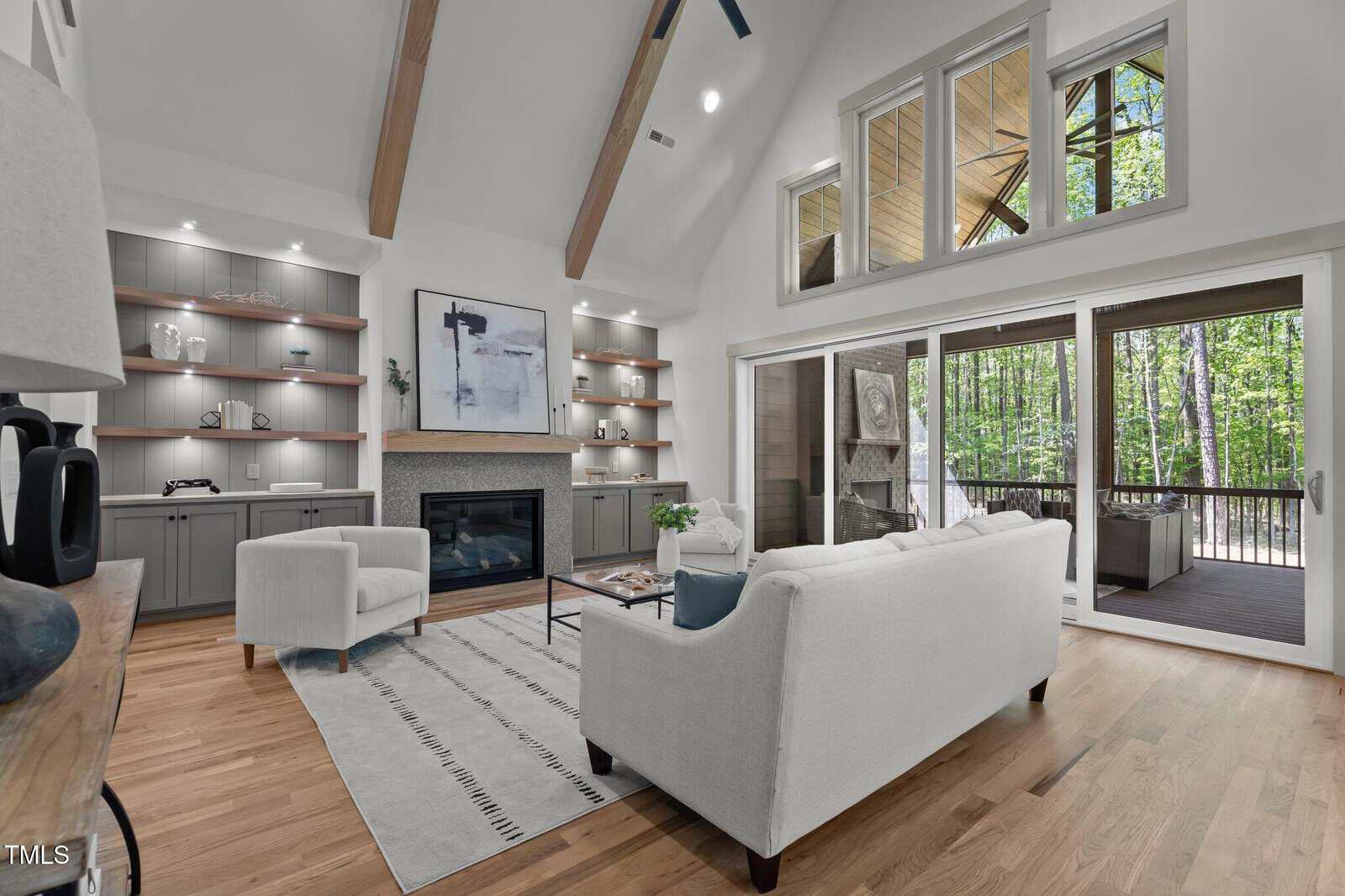 Modern living room with vaulted ceiling, wood beams, built-in shelves, fireplace, white sofas, large windows, and sliding doors opening to a covered porch overlooking a wooded area.