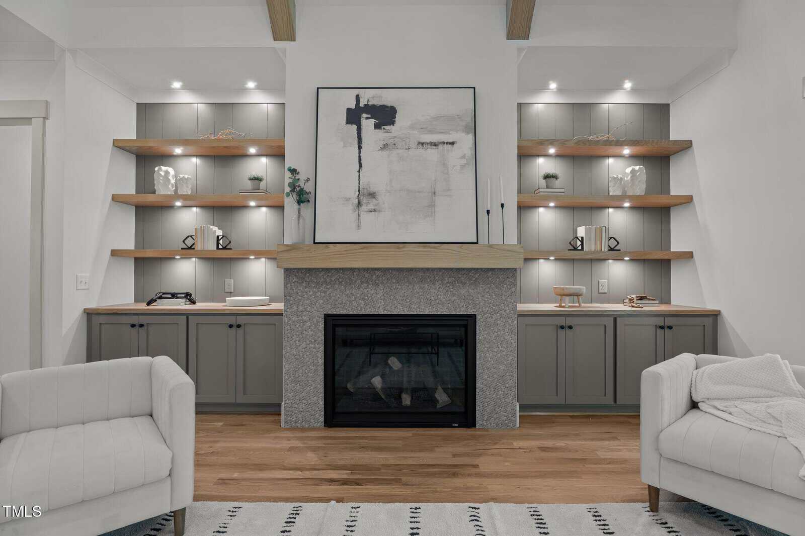 Modern living room with a gray stone fireplace, abstract art above, built-in shelves with decor, and closed cabinets. Two armchairs and a striped rug are visible in the foreground. Warm wooden floor and soft lighting create a cozy feel.