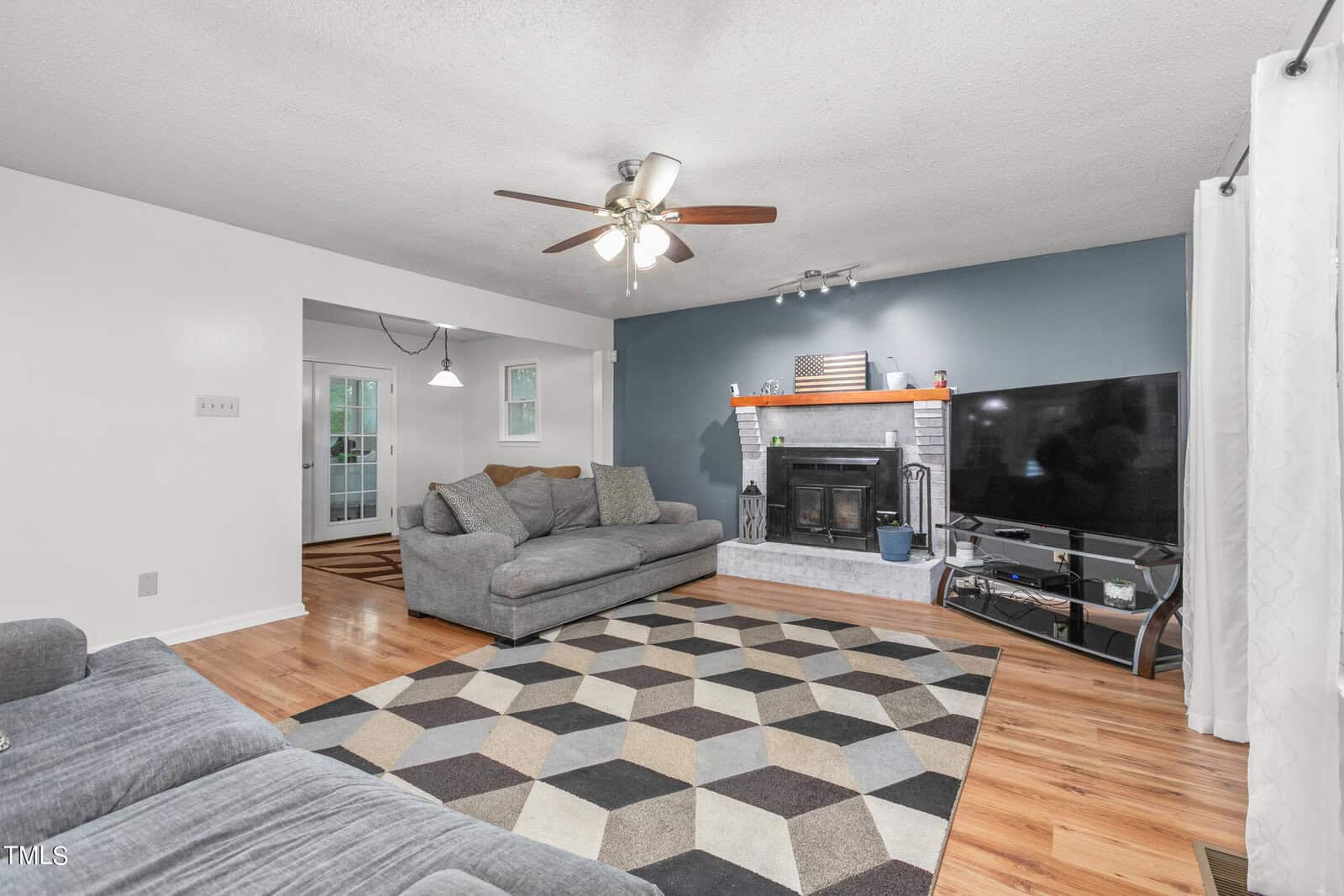 A cozy living room with a geometric-patterned rug, two grey sofas, a large flat-screen TV, and a brick fireplace. The walls are painted white and blue, and there's a ceiling fan with lights. Natural light comes in through a window with white curtains.