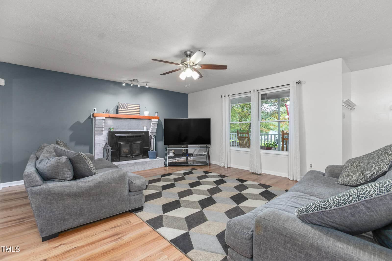 A cozy living room with a geometric-patterned rug, two gray couches, and a large flat-screen TV. A brick fireplace decorates the blue accent wall. Natural light streams through a window overlooking a green outdoor space. Ceiling fan and wooden floor.