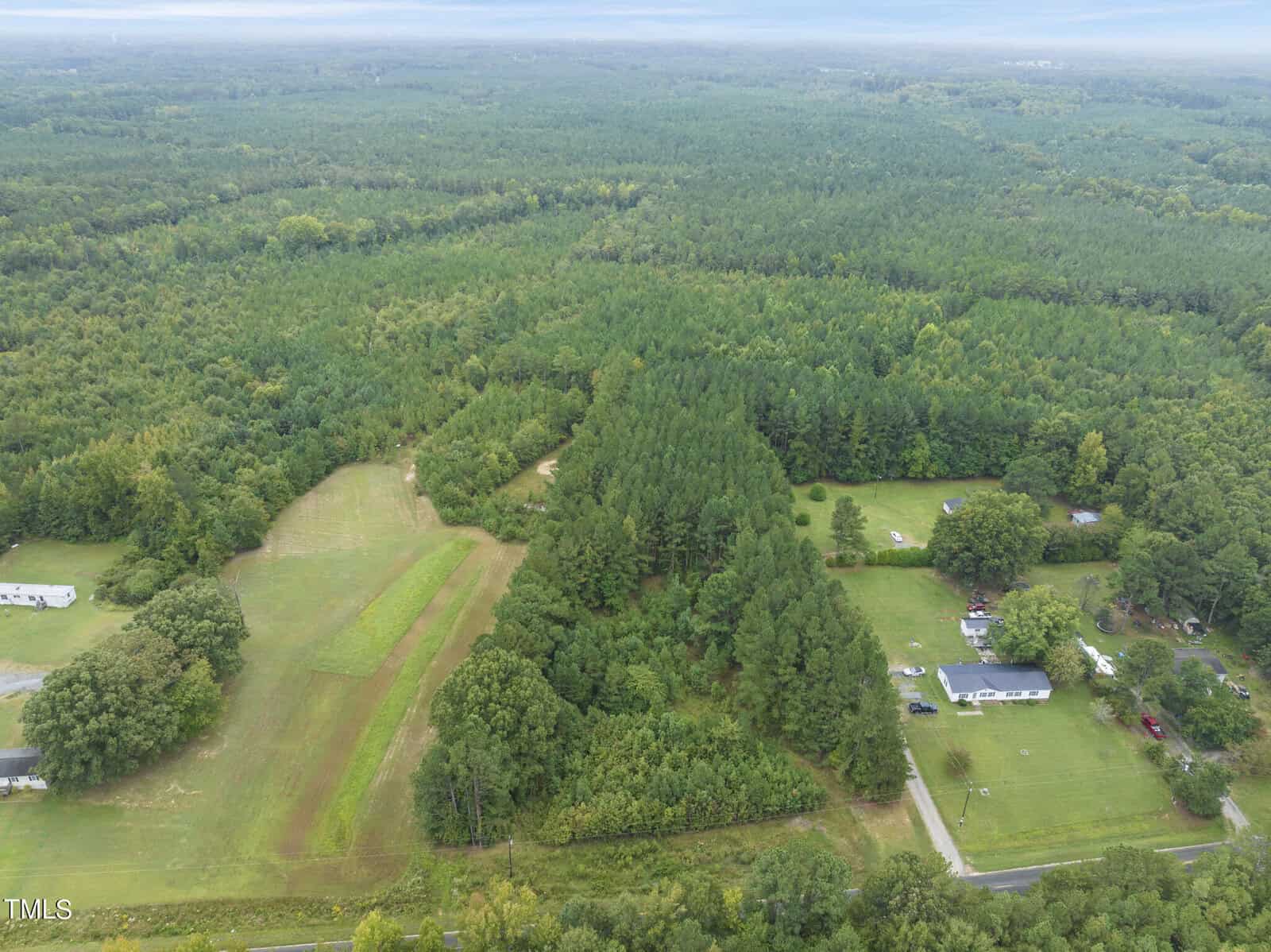 Aerial view of a rural landscape featuring dense green forests and patches of open land near Oxford. A few houses and cleared areas are visible among the trees, including one at 2602 Bodie Currin Road. A road runs horizontally across the bottom of the image, while the sky appears cloudy.