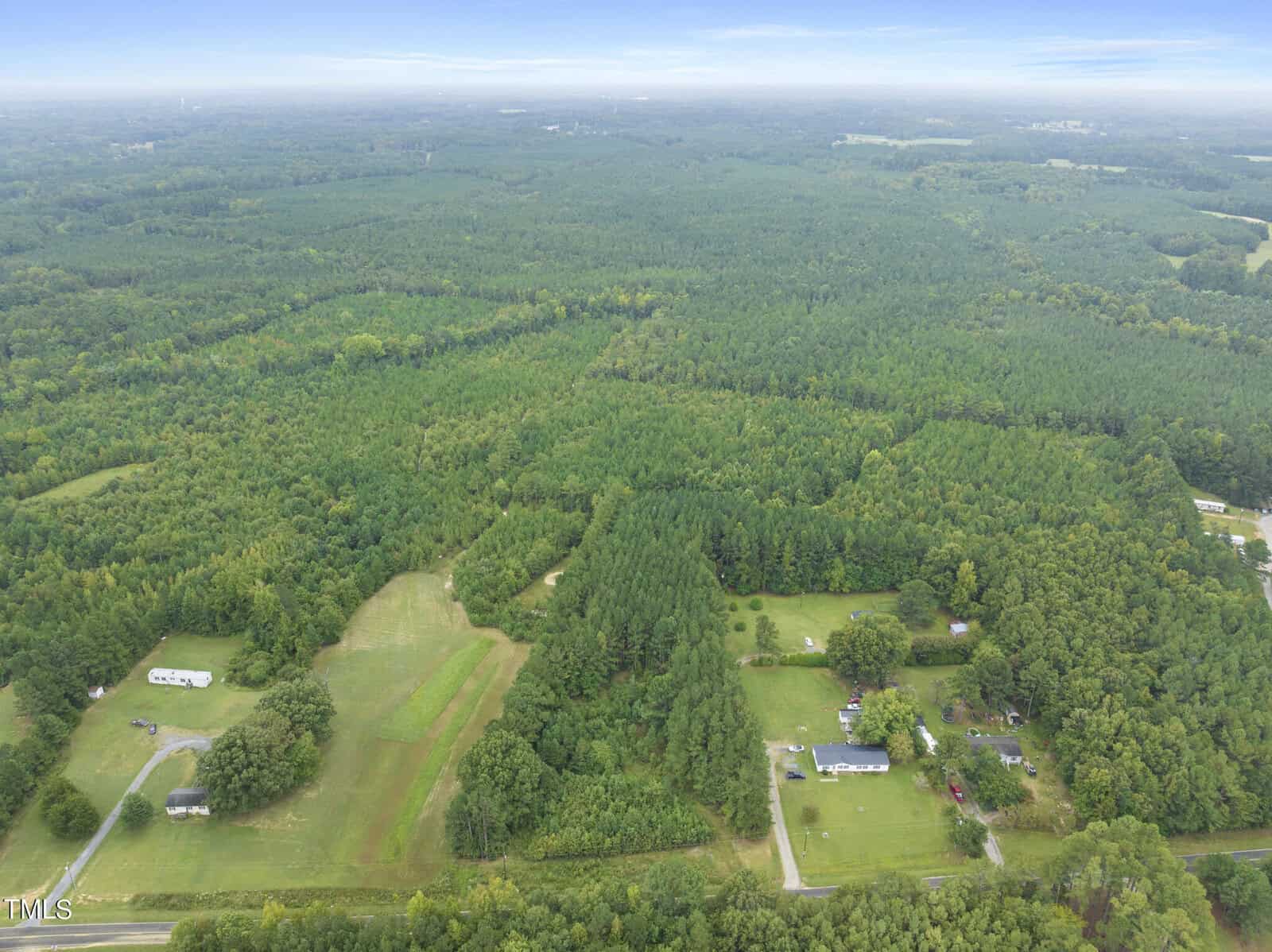 Aerial view of a vast, lush green forest with scattered clearings and a few houses. Patches of fields and tree-lined roads are visible, surrounded by expansive wooded areas under a blue sky with scattered clouds.