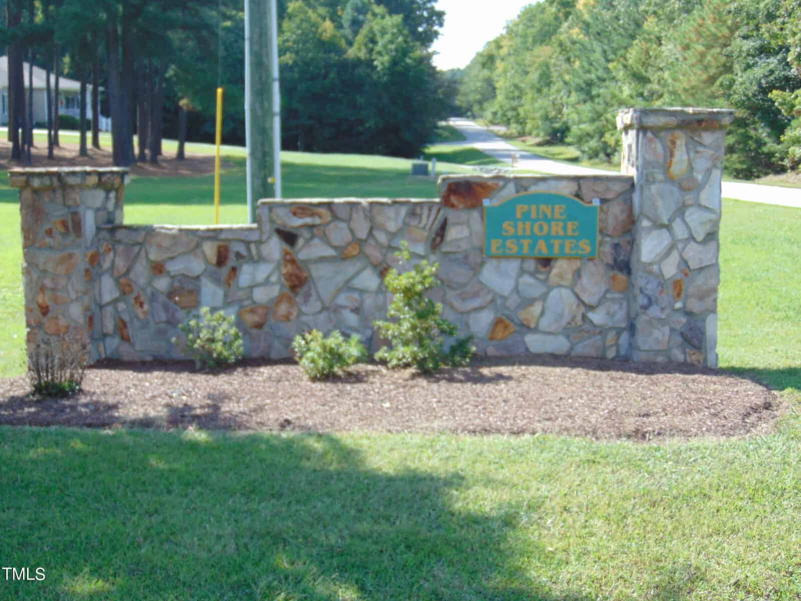 Stone sign for "Pine Shore Estates," surrounded by grass and bushes, with trees and a road in the background.
