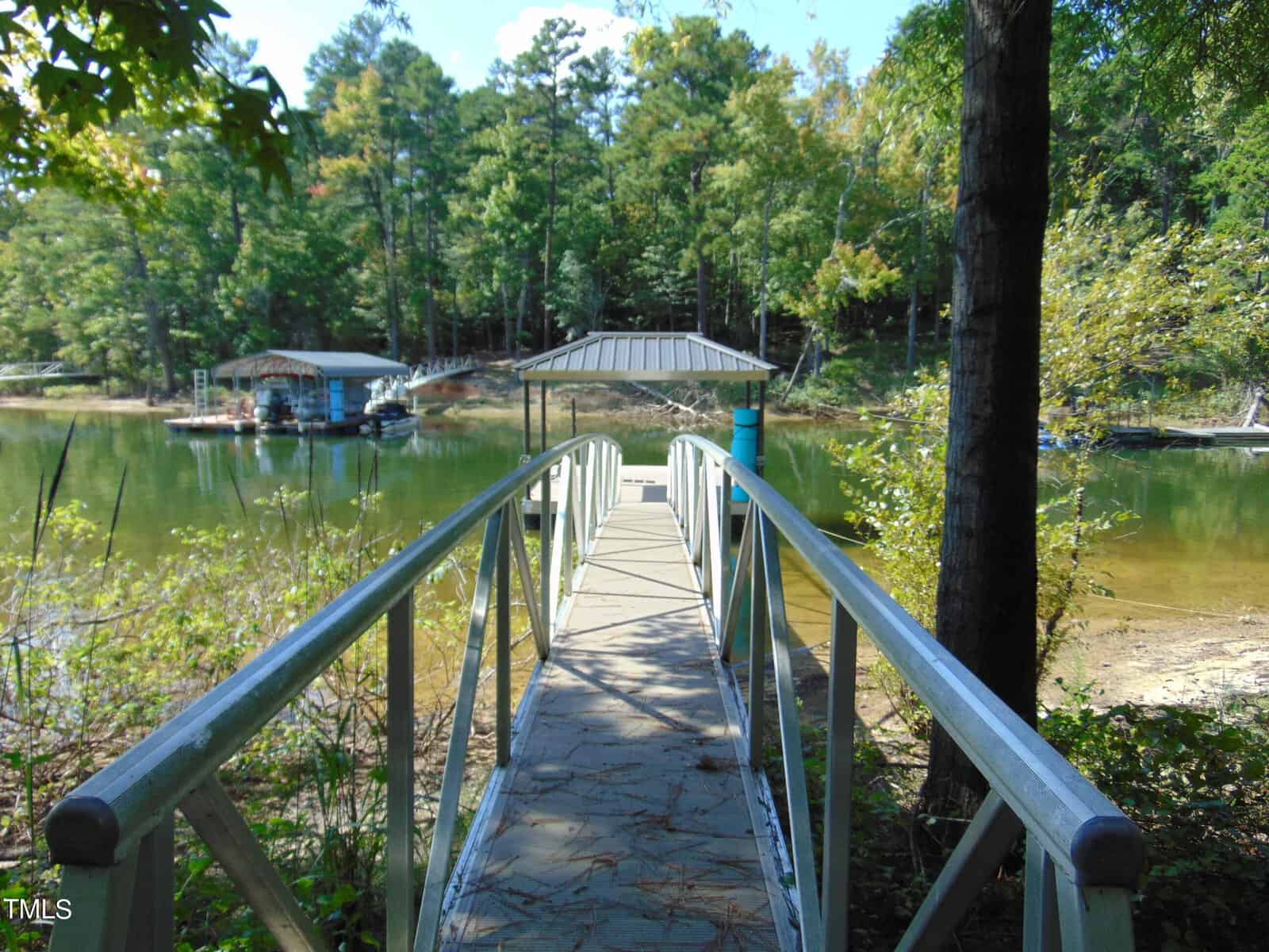 A narrow wooden bridge with railings leads to a small dock on a tranquil lake. Surrounding the lake are lush trees and a boat is visible to the left under a canopy. The scene is peaceful and surrounded by nature.