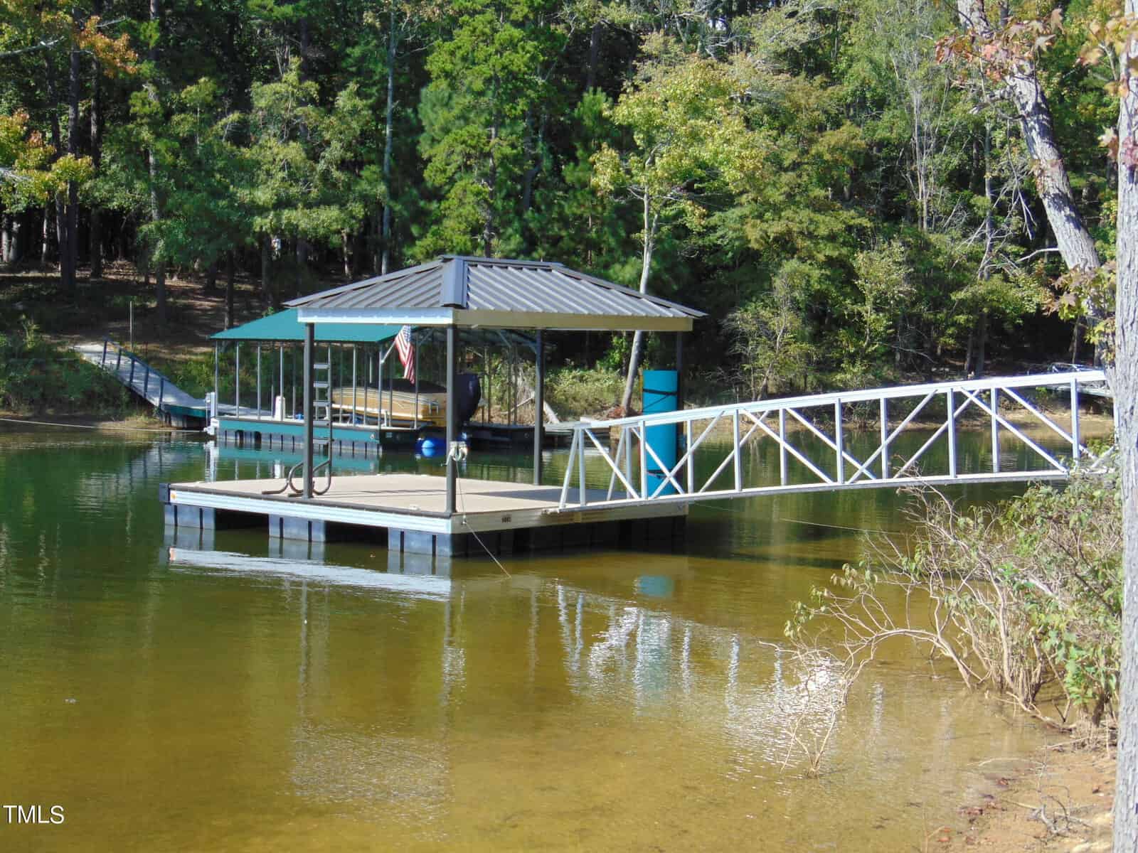 A small dock with a covered shelter extends into a calm lake, surrounded by lush green trees. A ramp connects the dock to the shore. The water is clear, revealing sandy areas beneath. A flag hangs from the shelter.