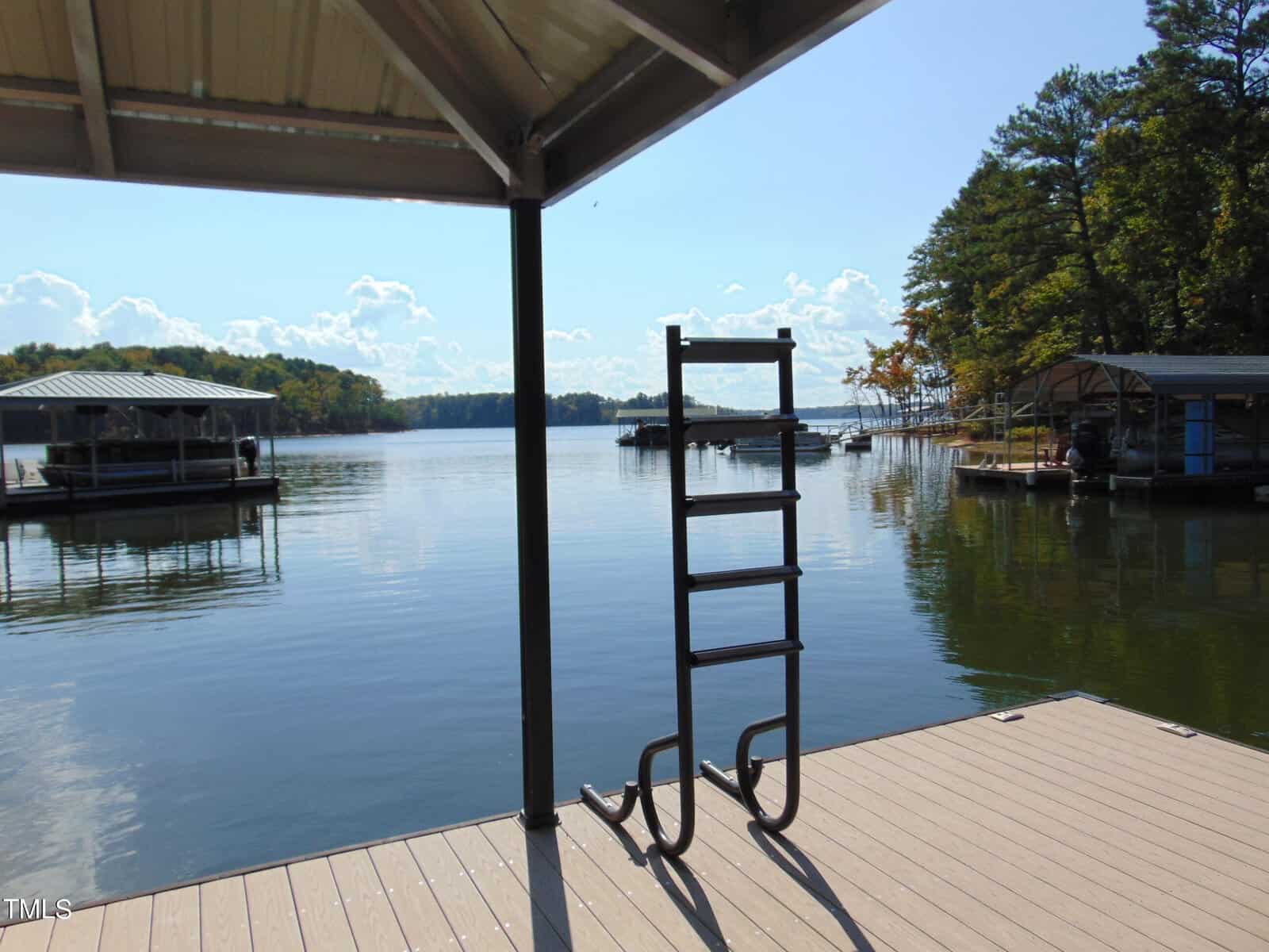 View from a dock with a metal ladder overlooking a calm lake. Several boat houses are visible in the distance, surrounded by trees under a clear blue sky.