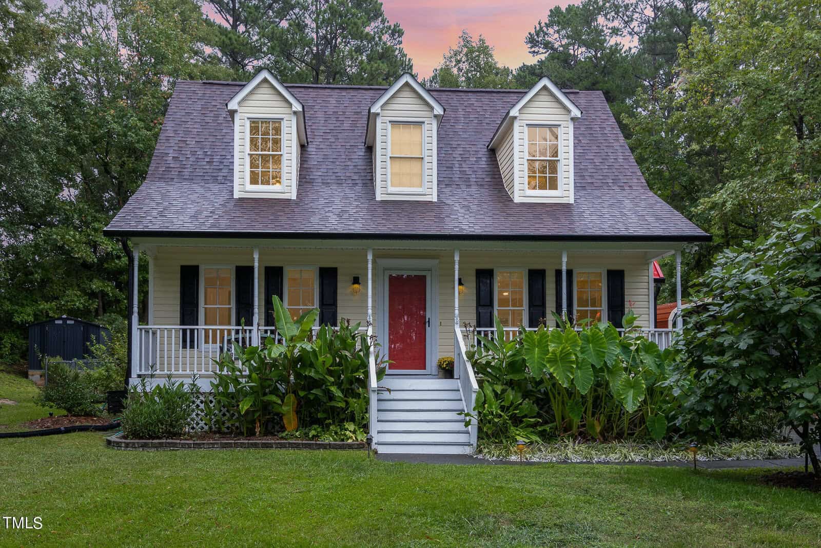 A charming two-story house with a front porch and three dormer windows. It has a red front door, white siding, and is surrounded by lush greenery and trees under a twilight sky.