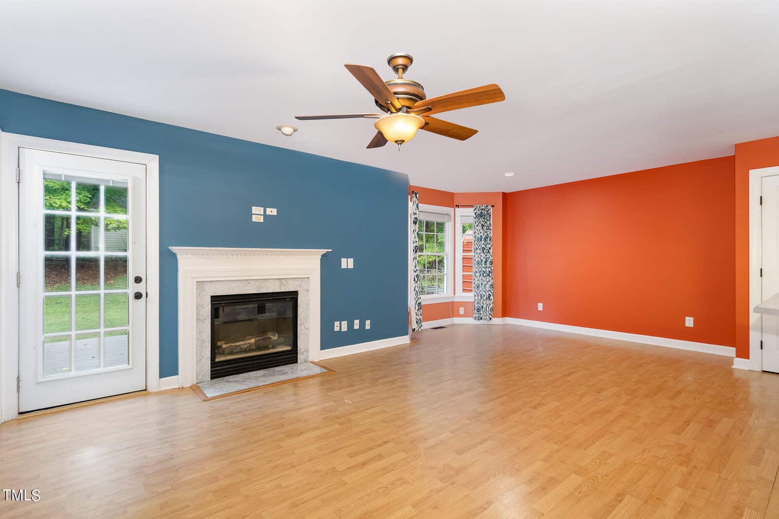 A modern living room with a blue and orange accent wall. The room features a ceiling fan, a fireplace, wooden flooring, and two large windows with patterned curtains. A glass door leads outside. The space is bright and spacious.