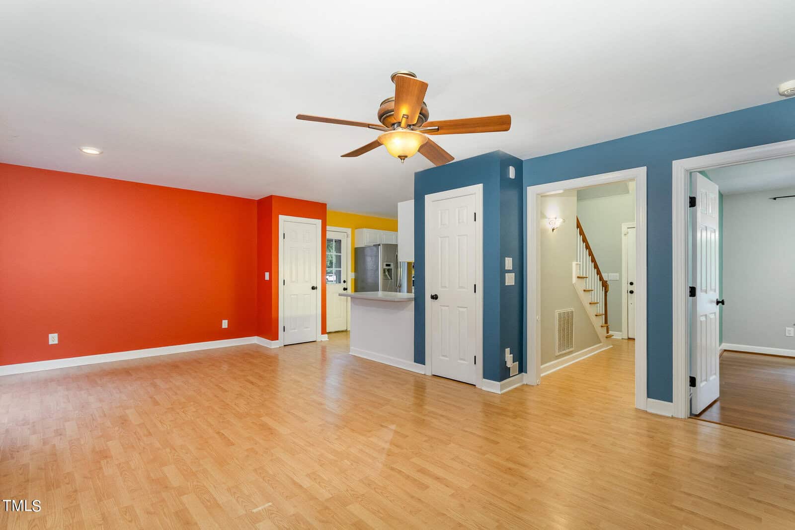 Spacious living room with light wood floors, vibrant red and blue accent walls, a ceiling fan, and multiple doorways leading to other rooms. A view of a staircase and open kitchen area in the background.