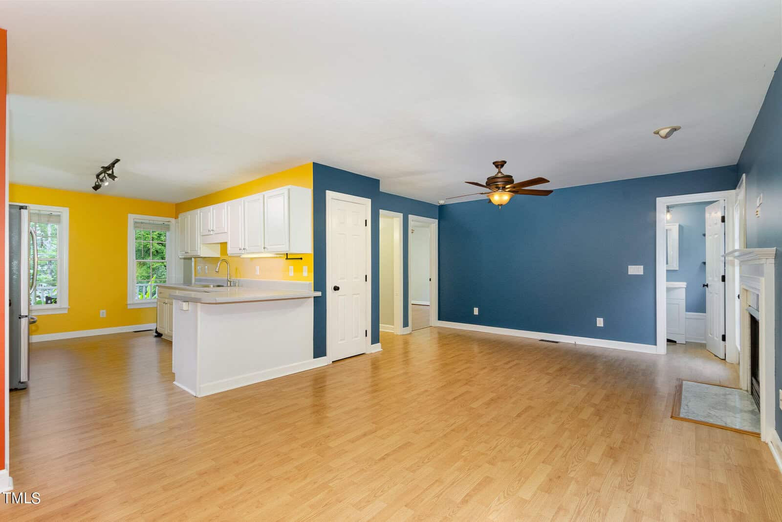 Spacious living room with blue and yellow walls, hardwood floors, and a ceiling fan. Open kitchen with white cabinets and countertops. A hallway leads to another room and a white door near the fireplace. Natural light from large windows.