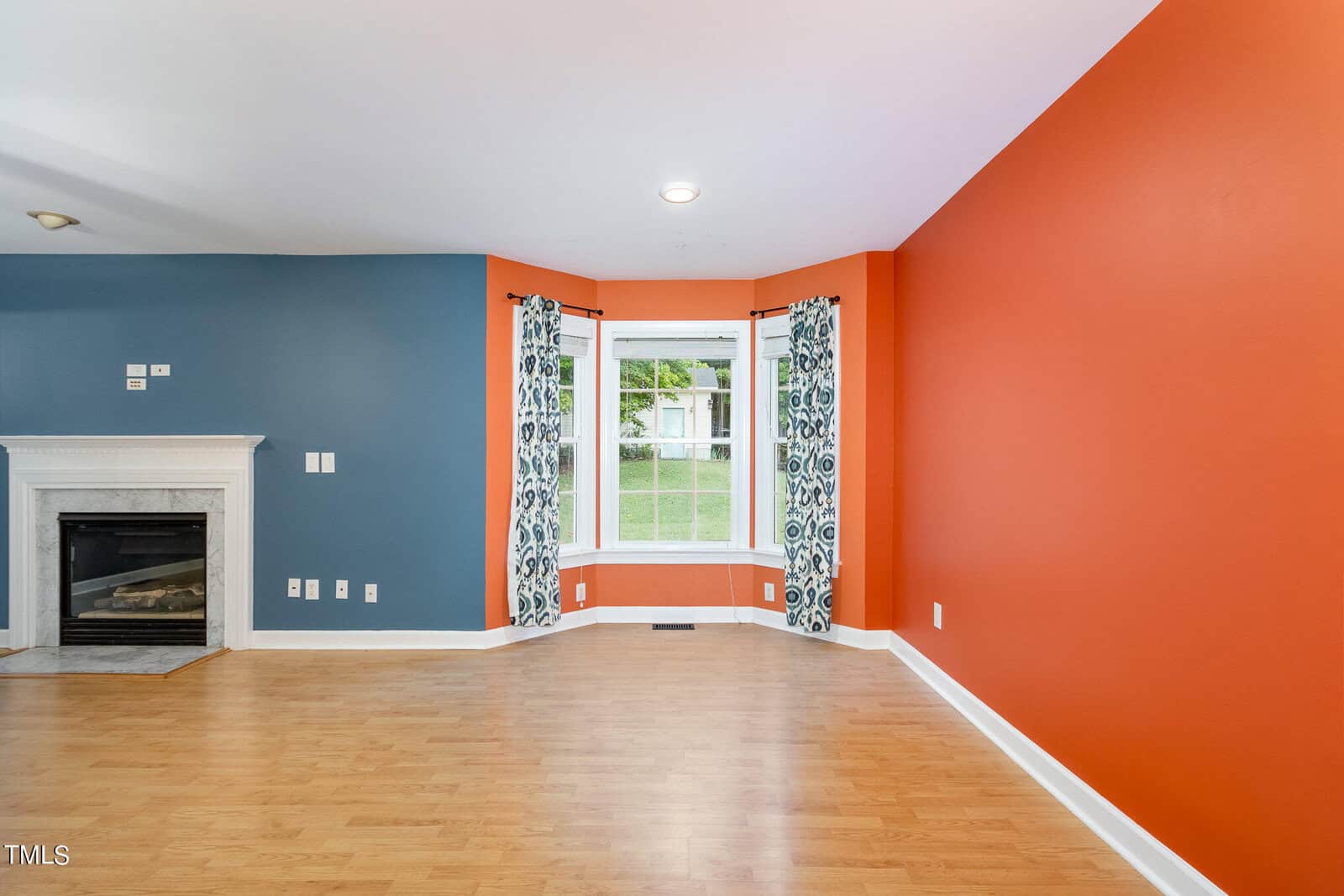 A living room with orange and blue walls, a white fireplace, and wooden flooring. A bay window features patterned curtains and offers a view of greenery outside.