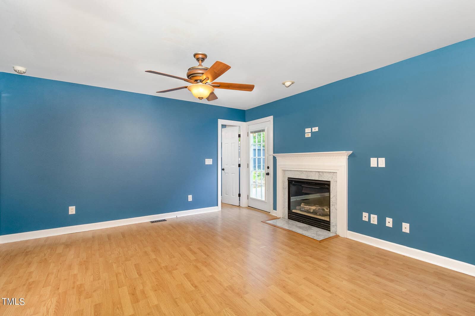 A bright, empty room with blue walls, wooden flooring, and a ceiling fan. A white fireplace is on the right wall, and a glass door opens to the outside. Electrical outlets and light switches are visible.