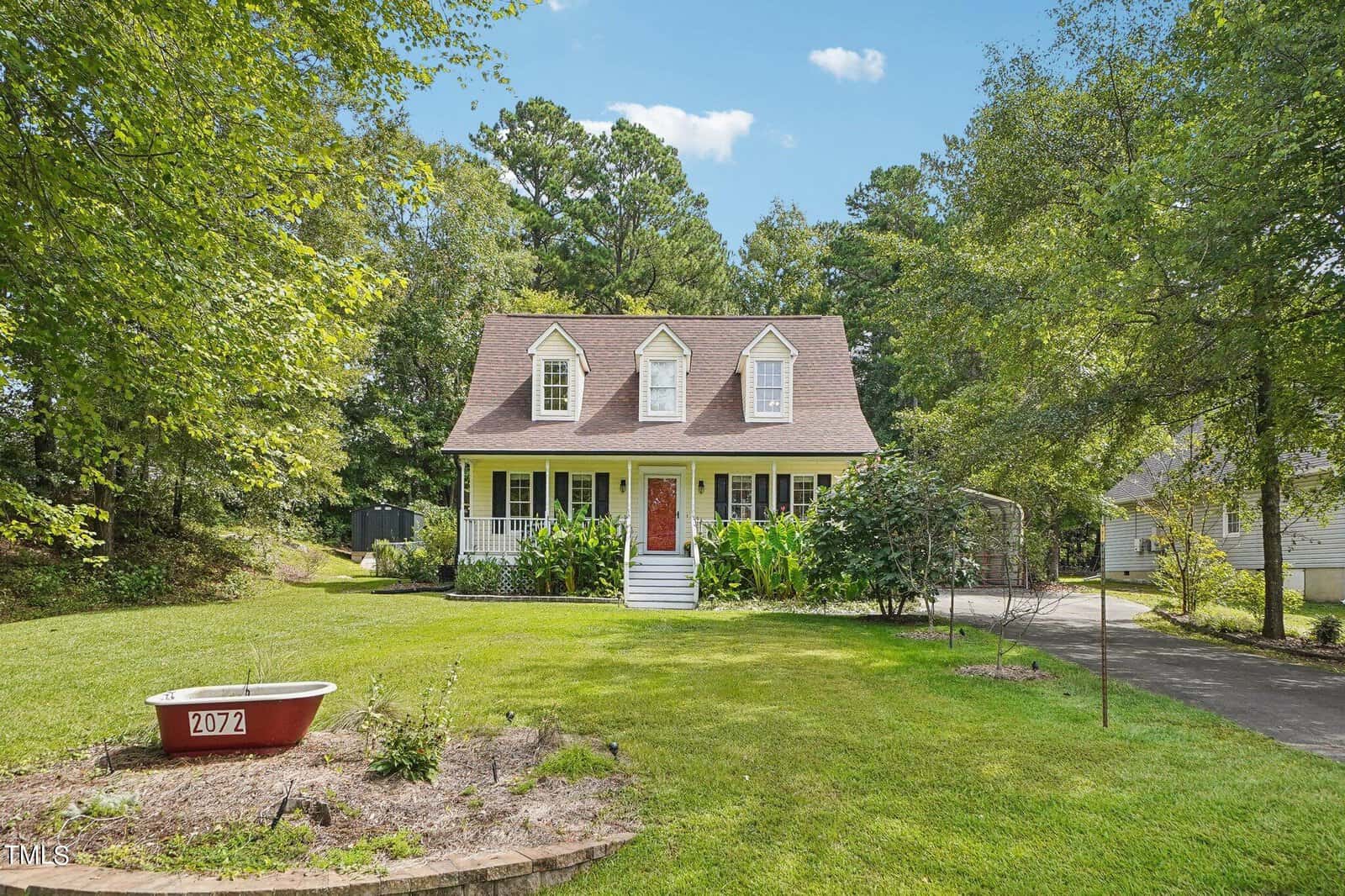 Located on 2072 Knight Street in Creedmoor, this charming two-story house boasts a steep gabled roof and three dormer windows. The red front door is framed by green shrubs and a manicured lawn. A paved driveway winds its way to a wooded area beneath the bright blue sky of a sunny day.