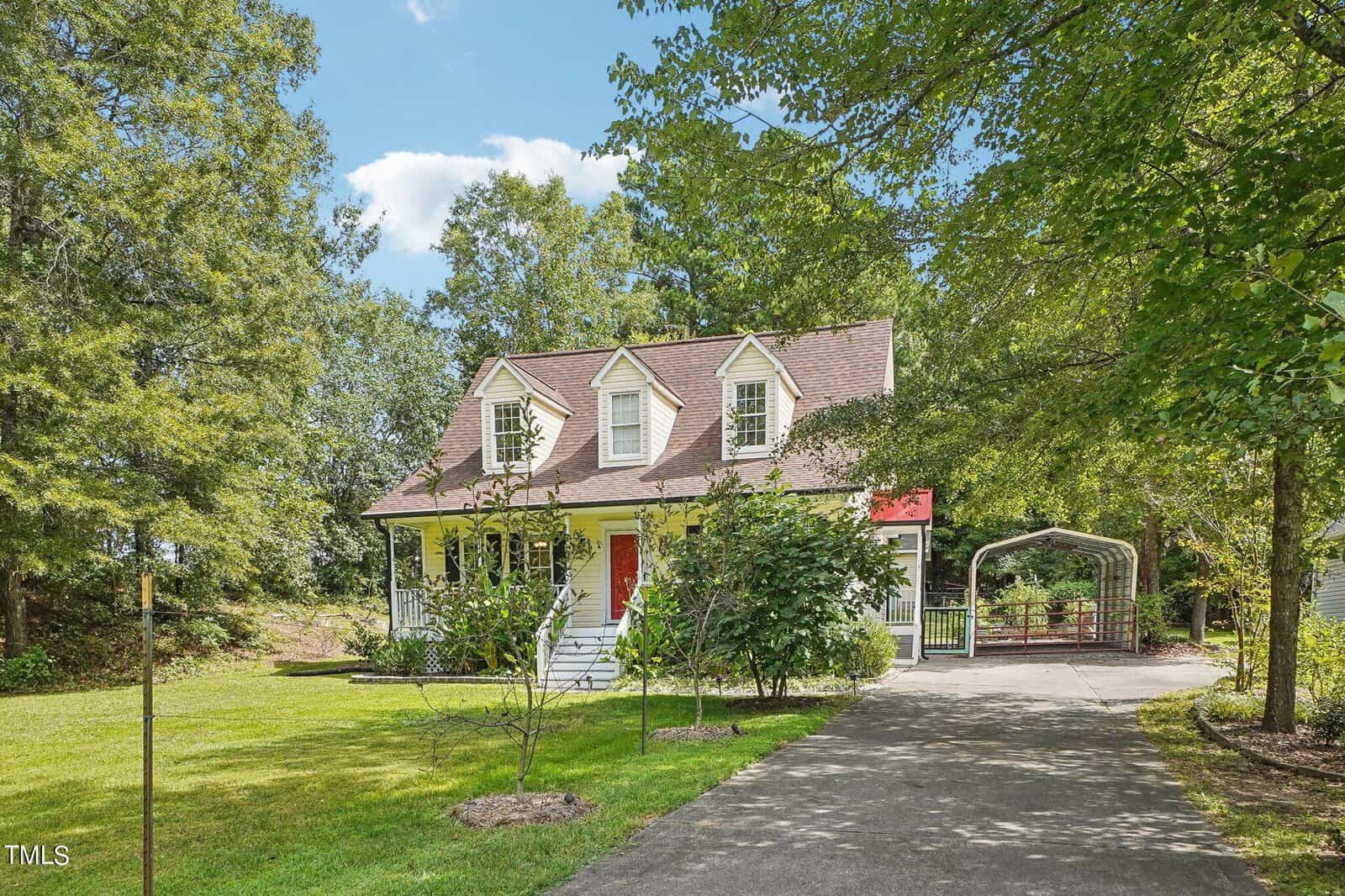A charming, small two-story house with dormer windows and a red door is surrounded by lush trees and a well-maintained lawn. A paved driveway leads to a carport on the right, while a clear blue sky is visible above.