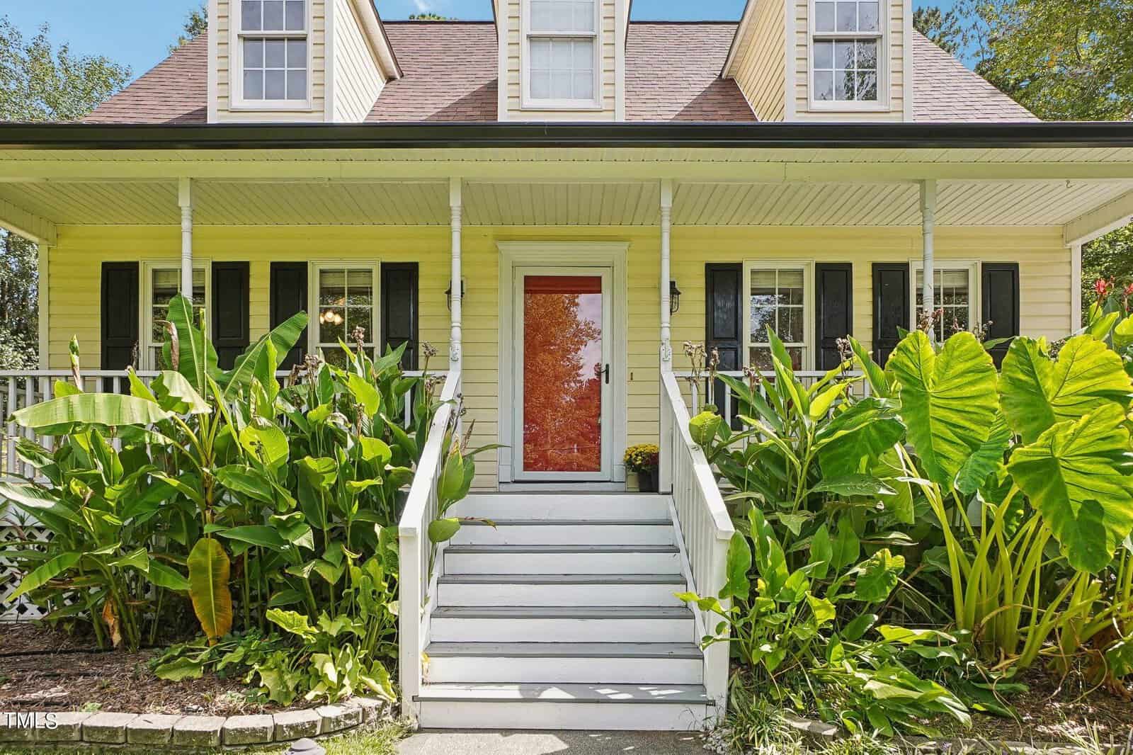 A charming house with a yellow exterior and a red front door. A white staircase leads to the entrance, flanked by lush green plants. The house has a covered porch with white railing and dormer windows on the roof. Trees are visible in the background.