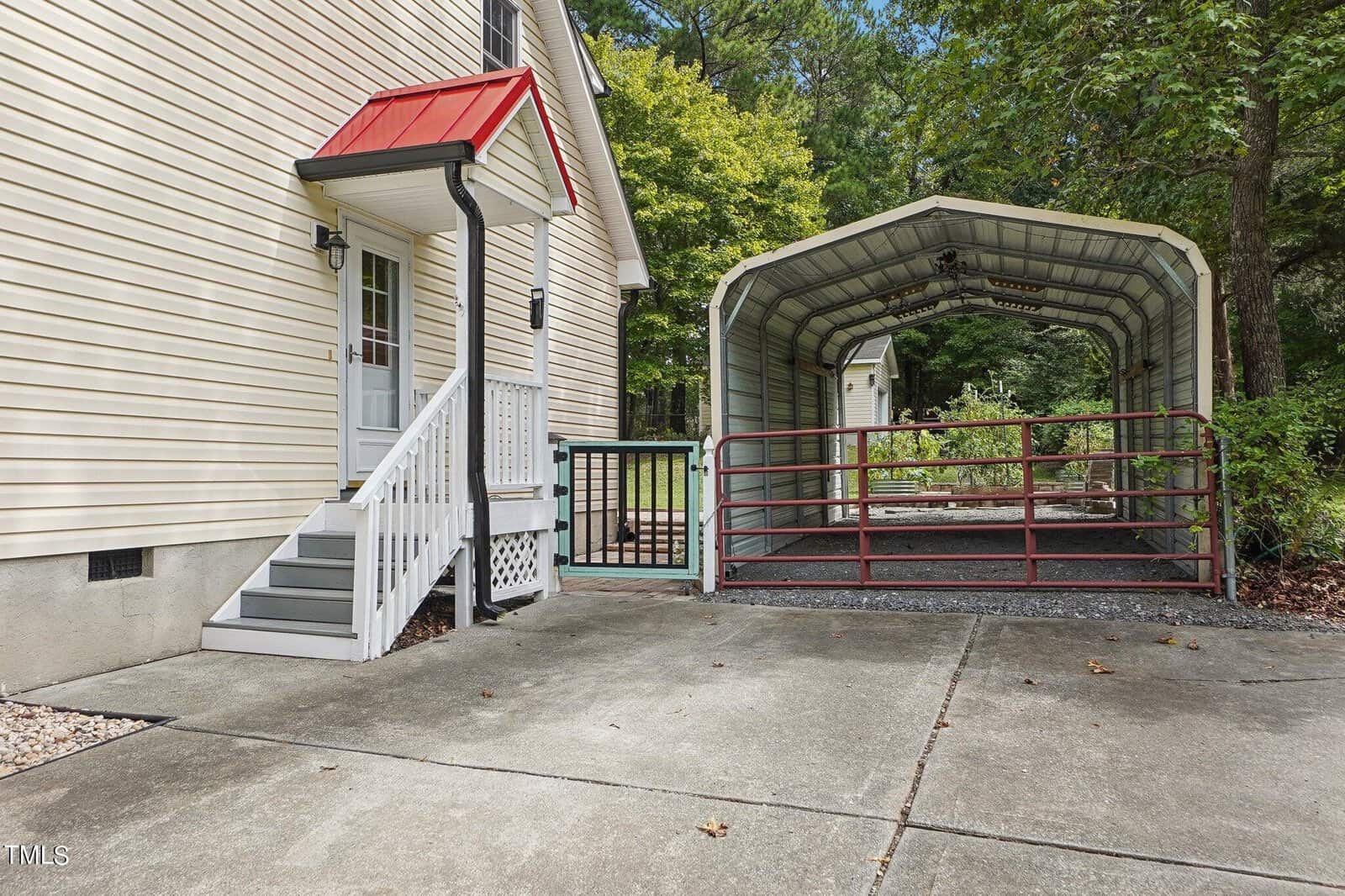 A single-story house with cream-colored siding and a red roof has a small set of steps leading to a door. Adjacent is a covered carport structure on a concrete driveway, surrounded by trees and greenery.