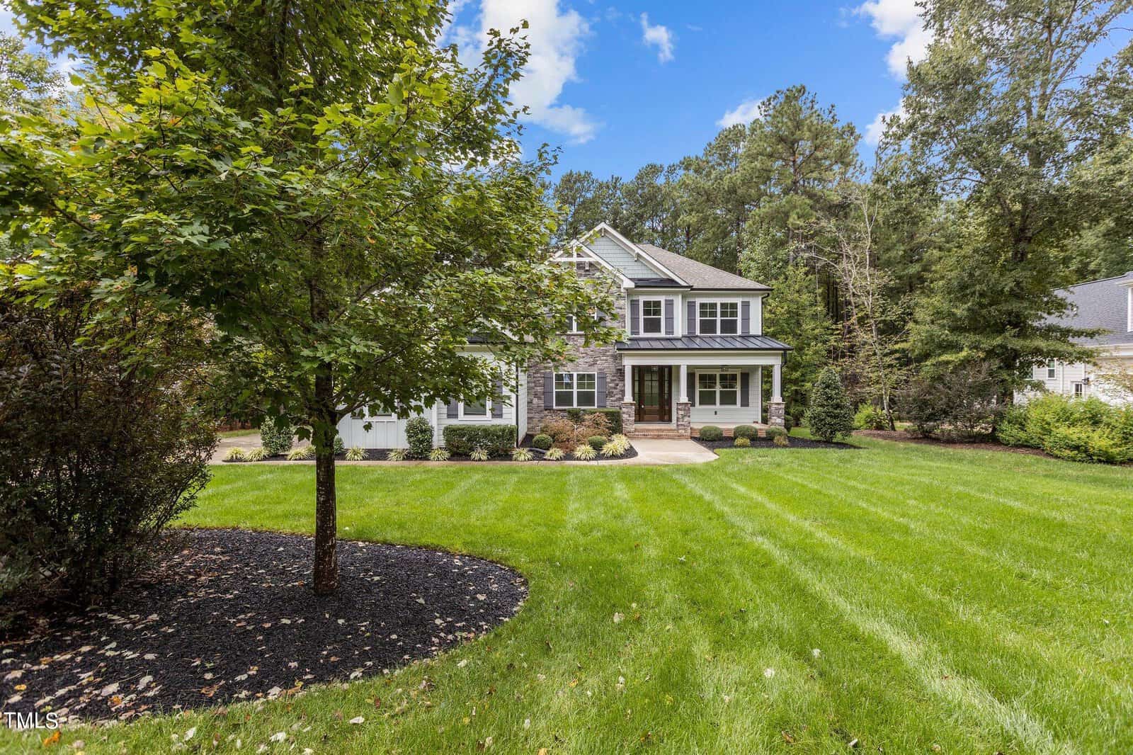A two-story house with a stone and siding exterior, surrounded by a lush green lawn and various trees. The sky is clear with a few clouds, and the setting appears peaceful and well-maintained.