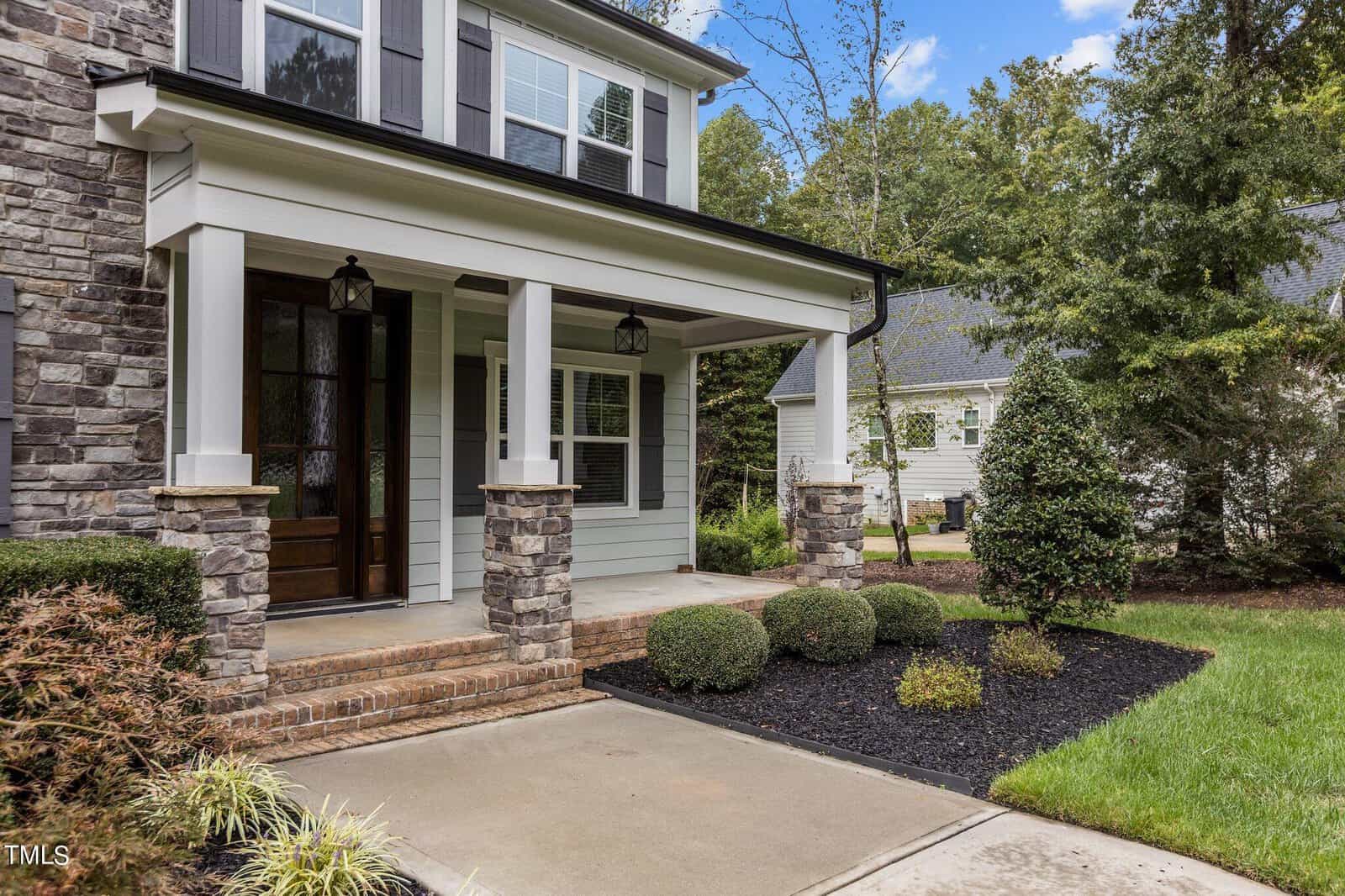 Front view of a house with stone and siding exterior, featuring a covered porch and dark wooden door. Neatly trimmed shrubs line the walkway, and trees are visible in the background against a clear blue sky.