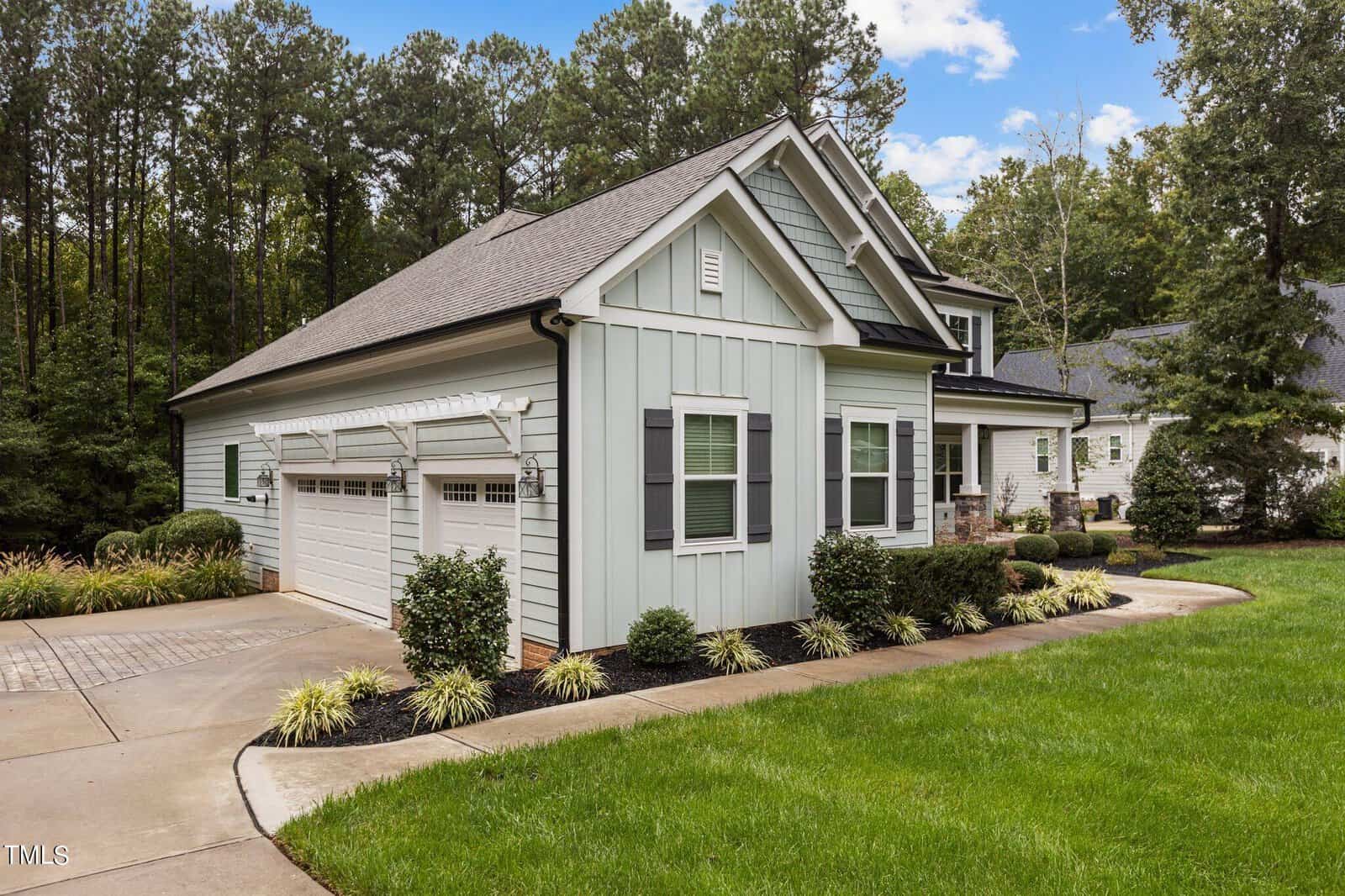 A modern suburban house with light gray siding, a dark roof, and white trim. It features a two-car garage, well-maintained bushes, a grassy lawn, and a backdrop of tall trees under a partly cloudy sky.