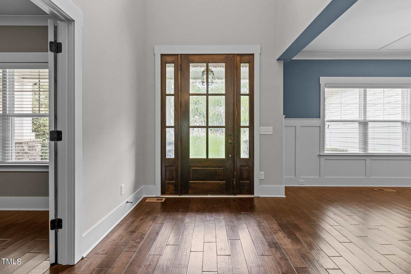 A bright foyer with a wooden front door featuring glass panels, opening into a room with hardwood floors. The walls are light gray with white trim, and a large window allows natural light into the space.