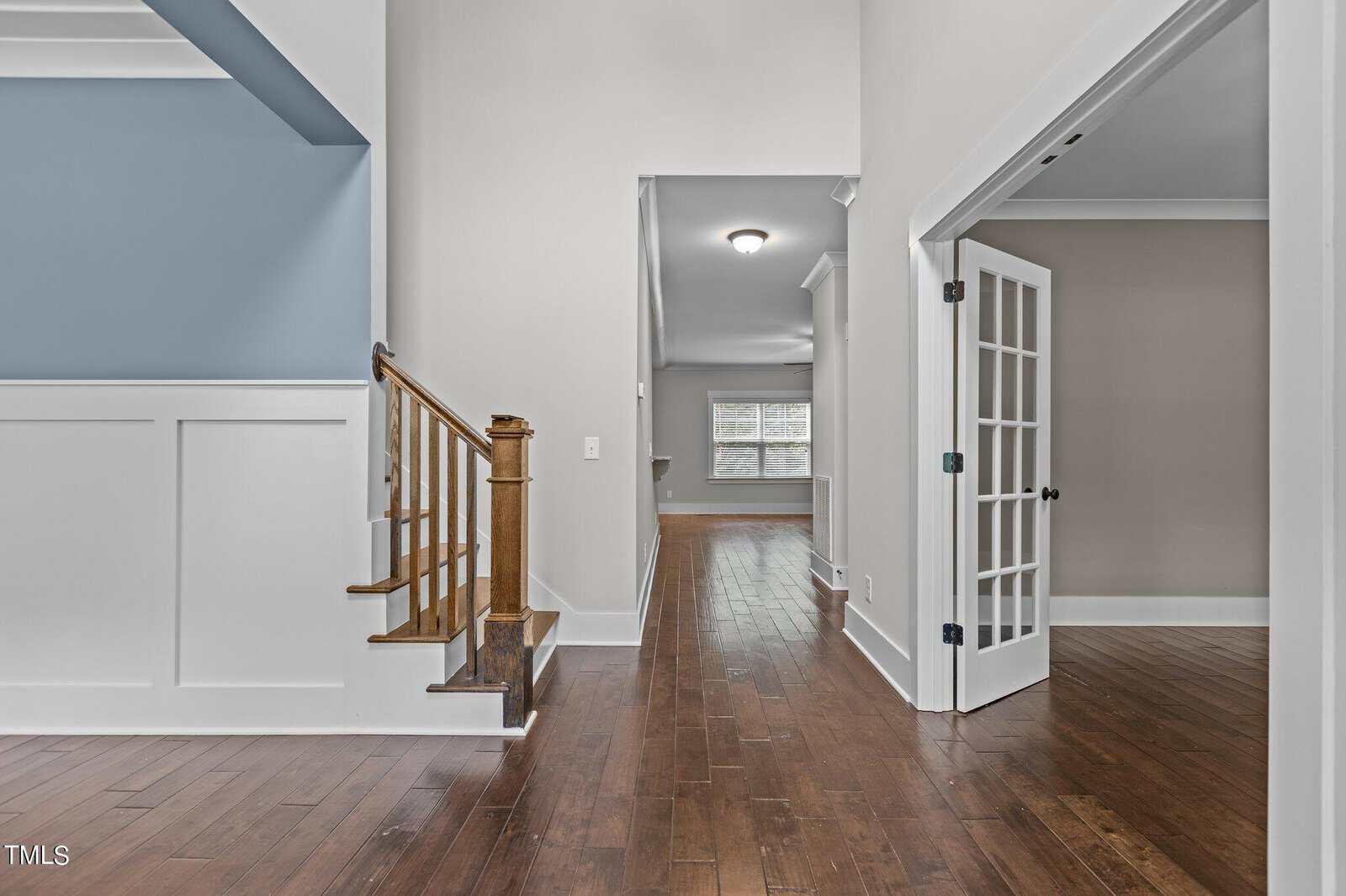 Interior of a modern home featuring a wooden staircase on the left, hardwood floors throughout, white walls, and a room with French doors on the right. A hallway leads to another room with large windows at the far end.