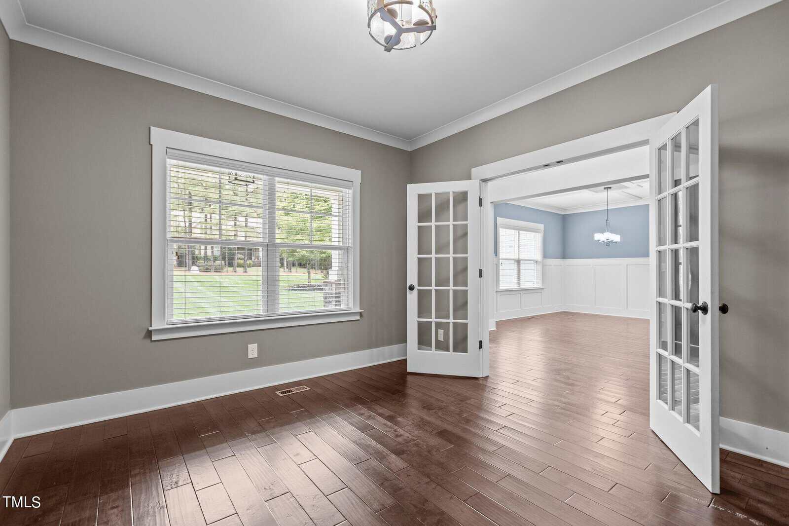 A spacious, empty room with light gray walls, hardwood floors, and a large window with blinds. French doors open to an adjacent room with similar design, both featuring modern overhead lights. Natural light fills the space, highlighting the clean interior.