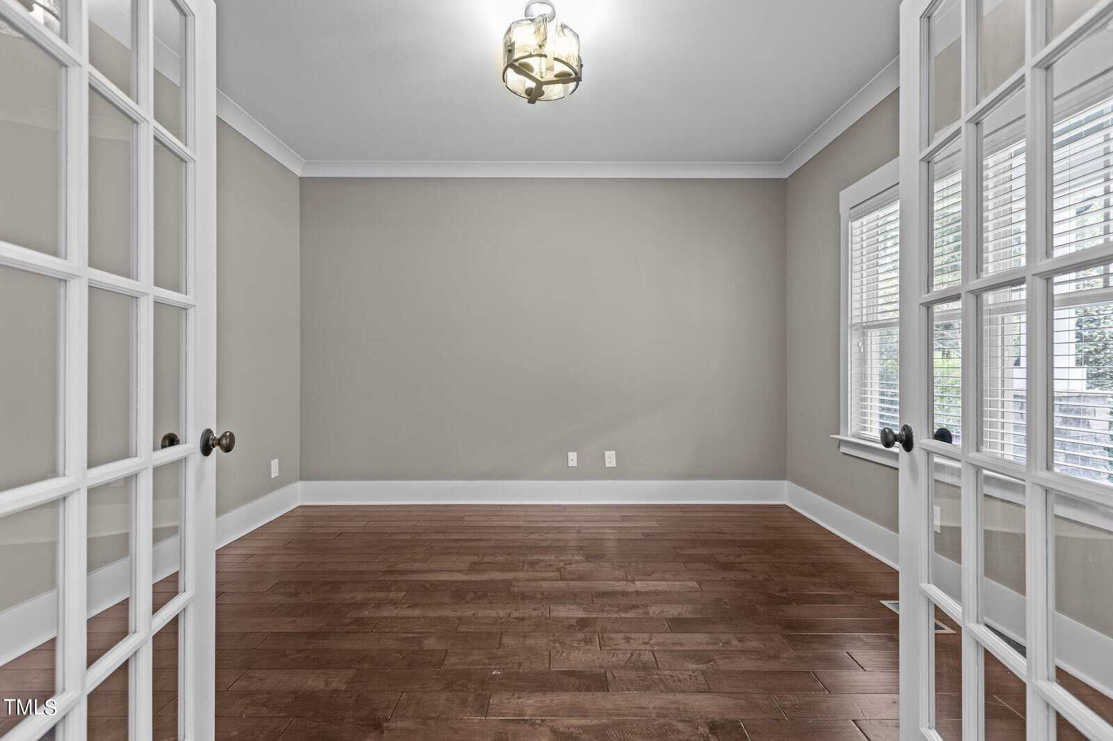 An empty room with light gray walls and dark hardwood floors, featuring a modern hanging light fixture. The room has French doors and two windows on the right, allowing natural light to enter.
