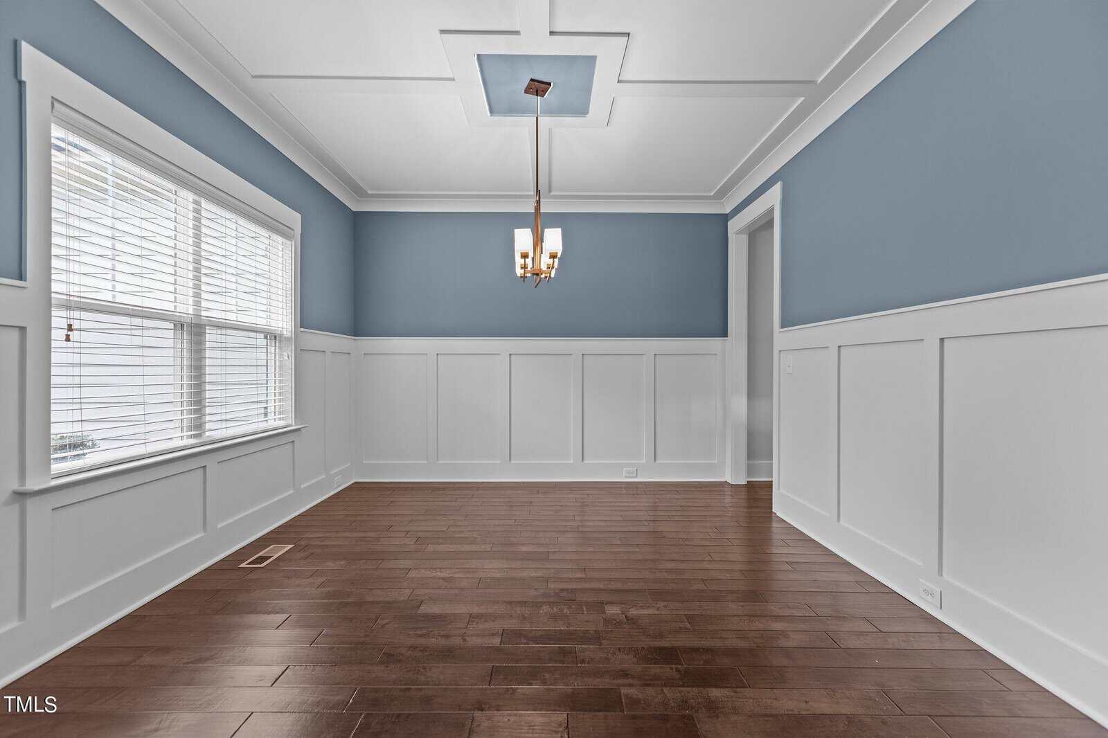 A spacious dining room with blue and white wainscoted walls, a hardwood floor, and a simple ceiling light fixture. A large window on the left lets in natural light.