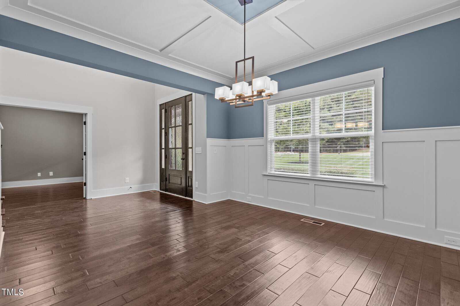 A modern dining room featuring light blue walls with white wainscoting. It has a dark wood floor and a white coffered ceiling with a rectangular chandelier. Large windows allow natural light to fill the space.