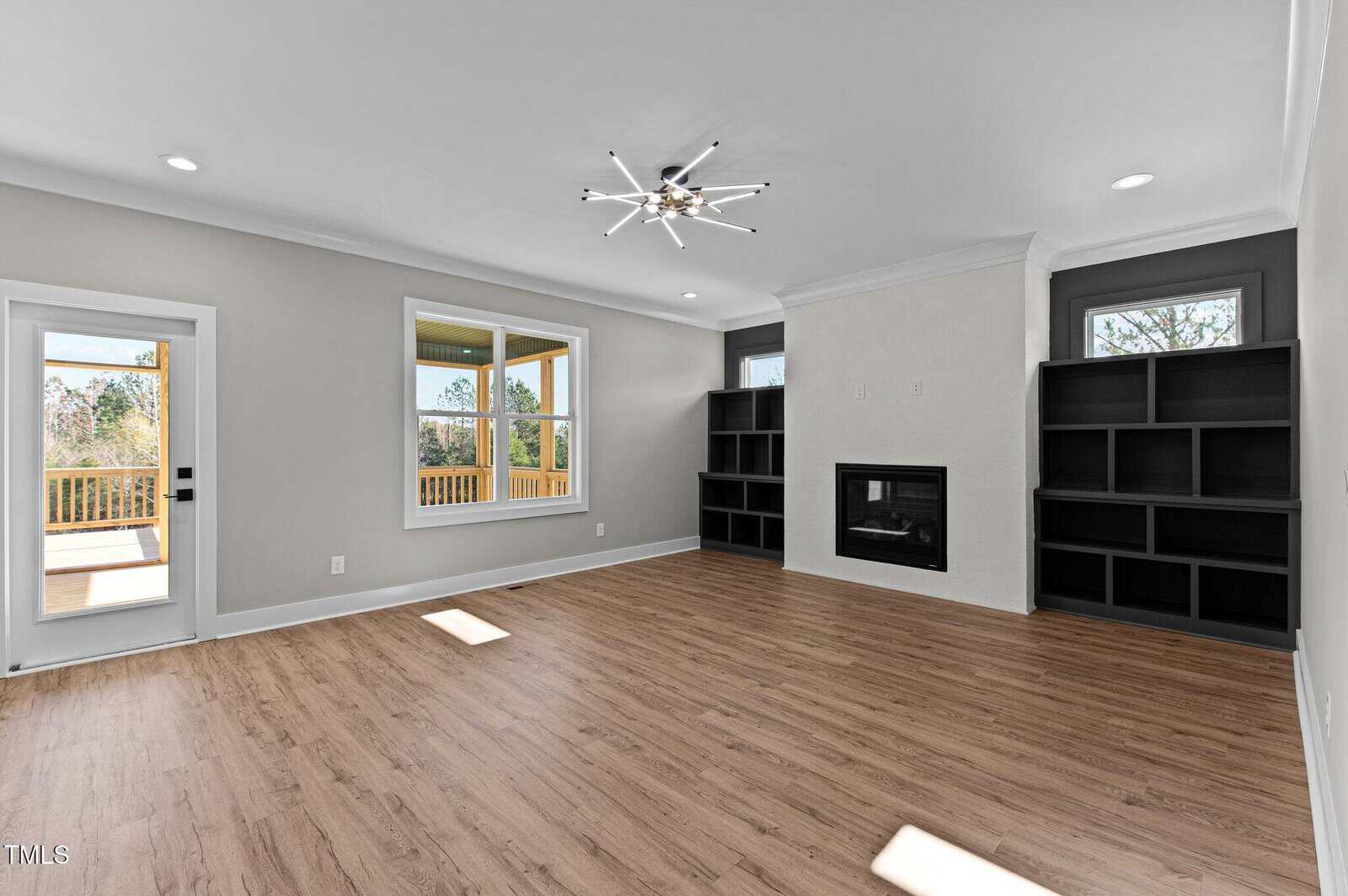 A modern living room with light wood flooring, white walls, black built-in shelves flanking a fireplace, recessed lighting, a contemporary ceiling light fixture, and large windows facing a deck.