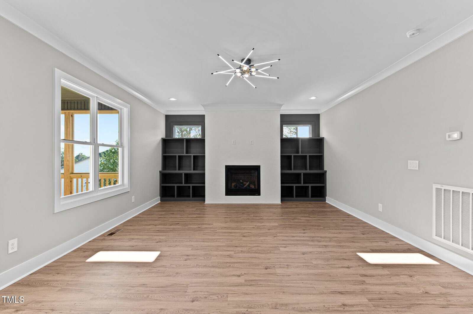 A bright living room with light wood flooring, light gray walls, a modern chandelier, a central fireplace, two built-in black shelving units, and windows on either side letting in natural light.