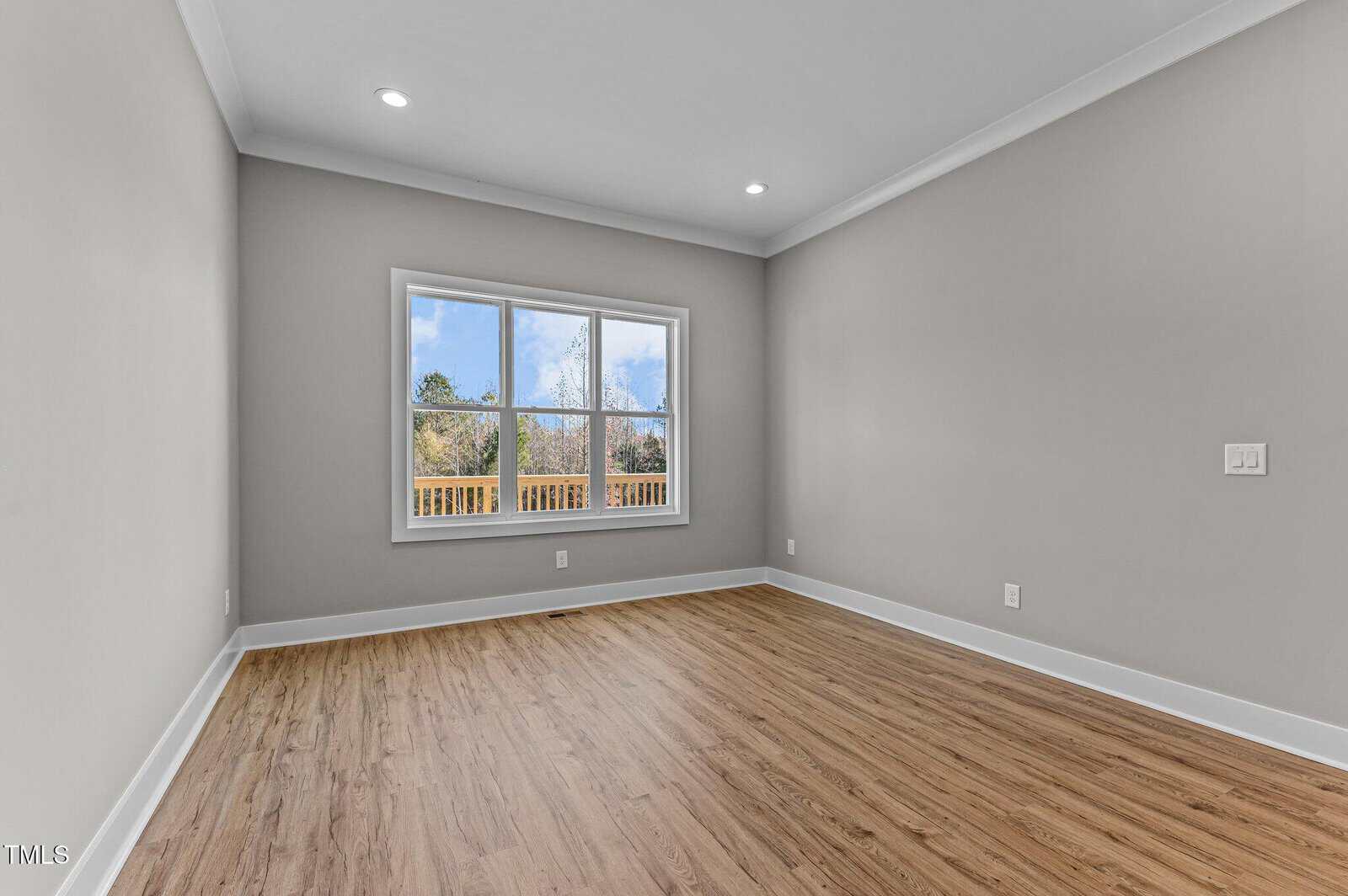 An empty room with light brown wood flooring, light gray walls, white trim, recessed ceiling lights, and a large window showing trees and blue sky outside.