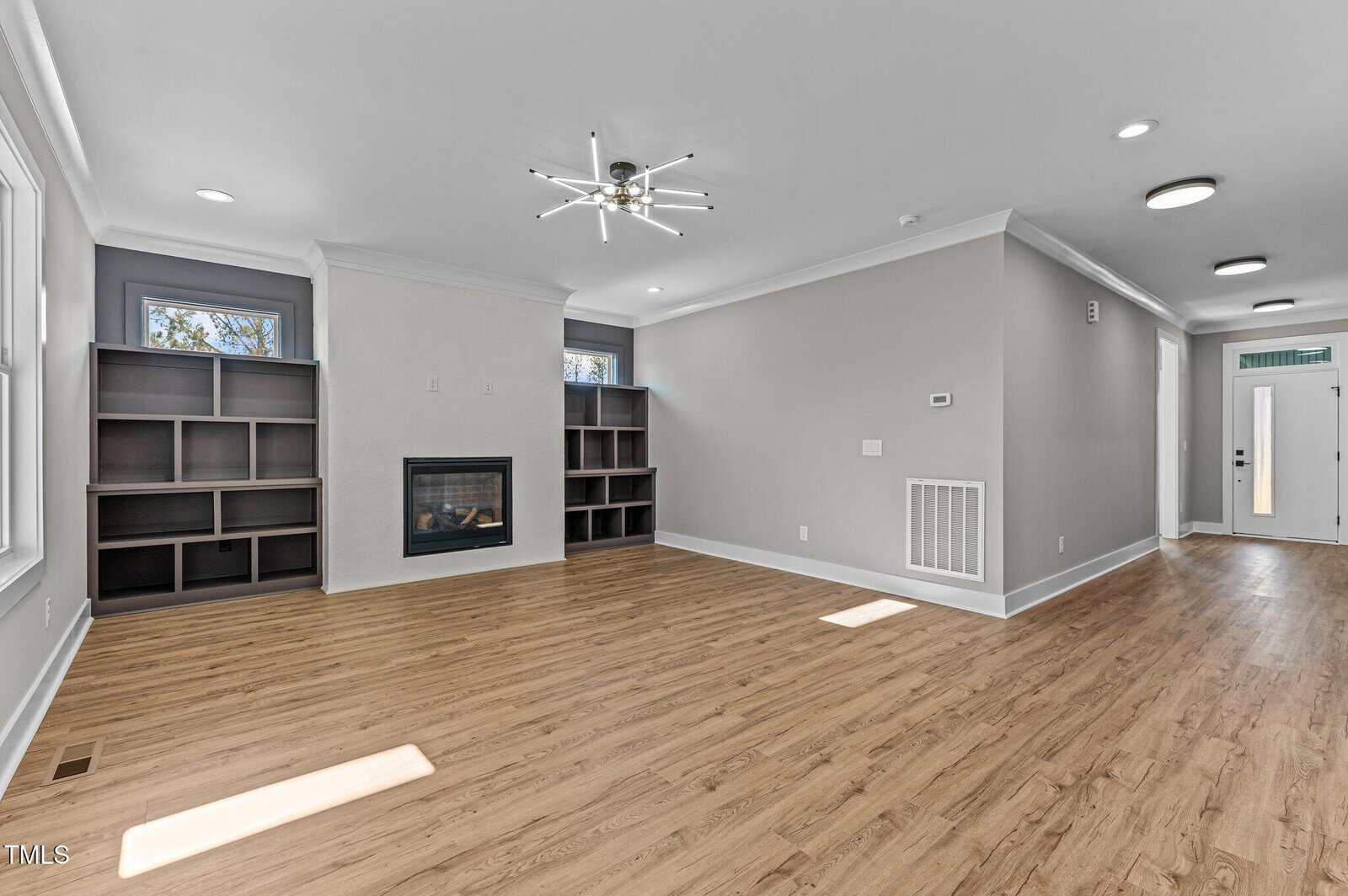 A spacious living room with light wood flooring, built-in shelves on either side of a modern fireplace, gray walls, recessed lighting, and a contemporary ceiling light fixture. A hallway with a front door is visible.
