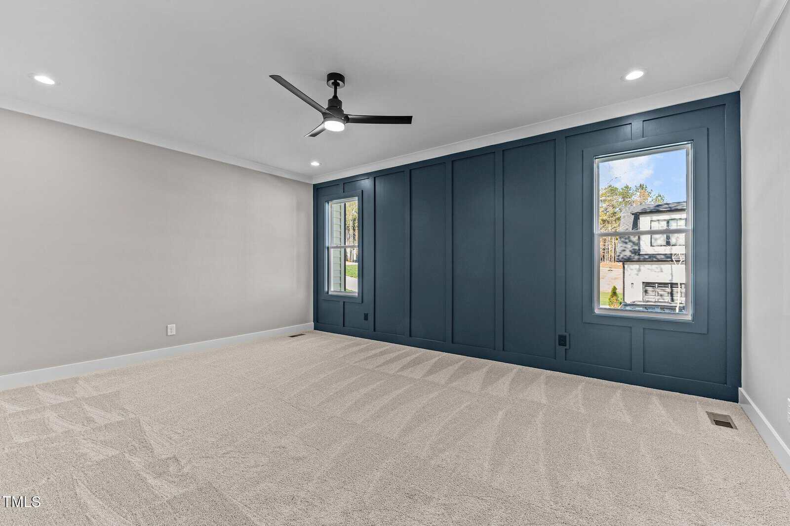 A spacious, empty room with beige carpet, light gray walls, a dark blue accent wall with two windows, a black ceiling fan, and recessed ceiling lights. Daylight is visible through the windows.