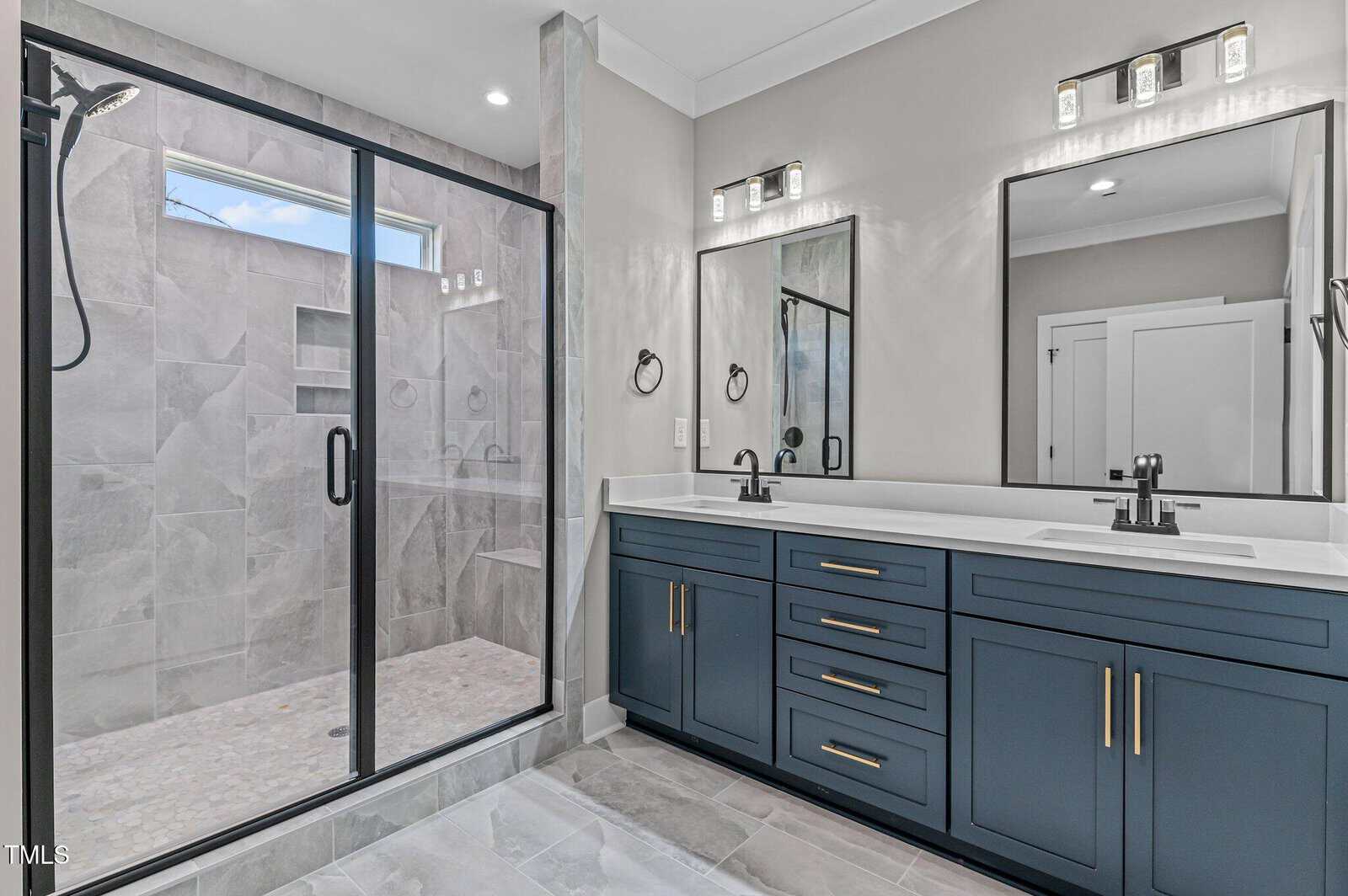 Modern bathroom with a large glass-enclosed shower, dual sinks with navy blue cabinets, black faucets, and two mirrors under sleek rectangular lights. Light gray tiles and walls create a bright, clean look.