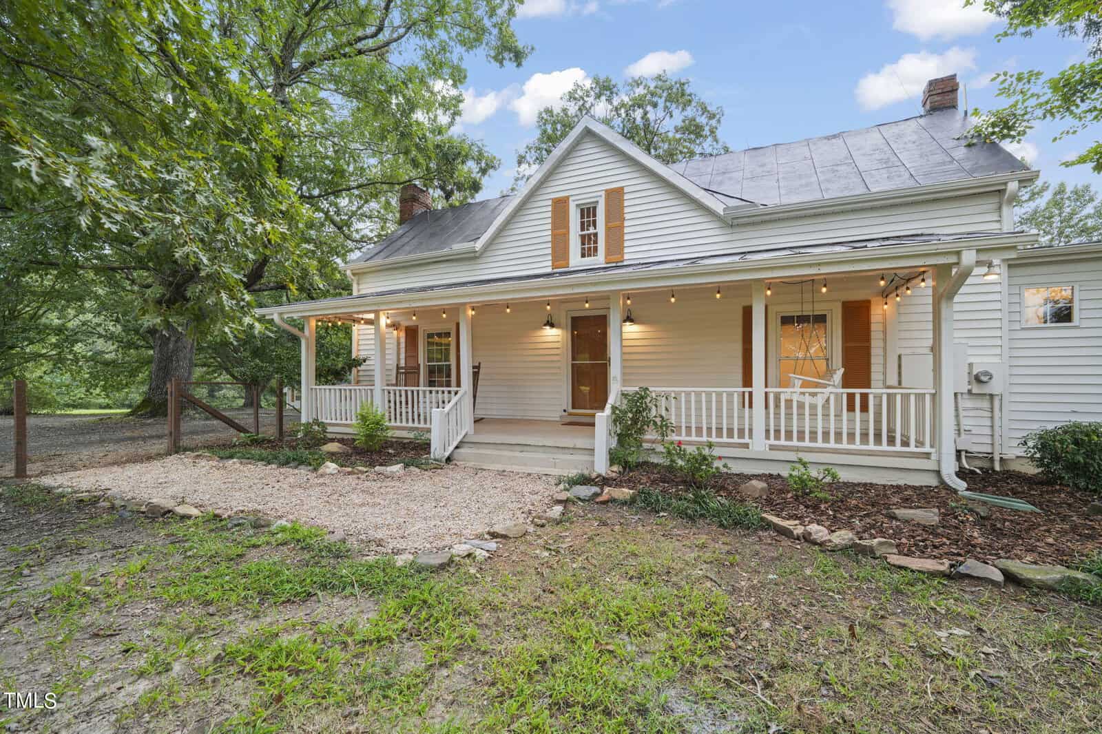 A charming white house with a wrap-around porch adorned with string lights. Surrounded by lush greenery and trees, the house features a gravel path leading to the entrance and wooden rocking chairs on the porch.
