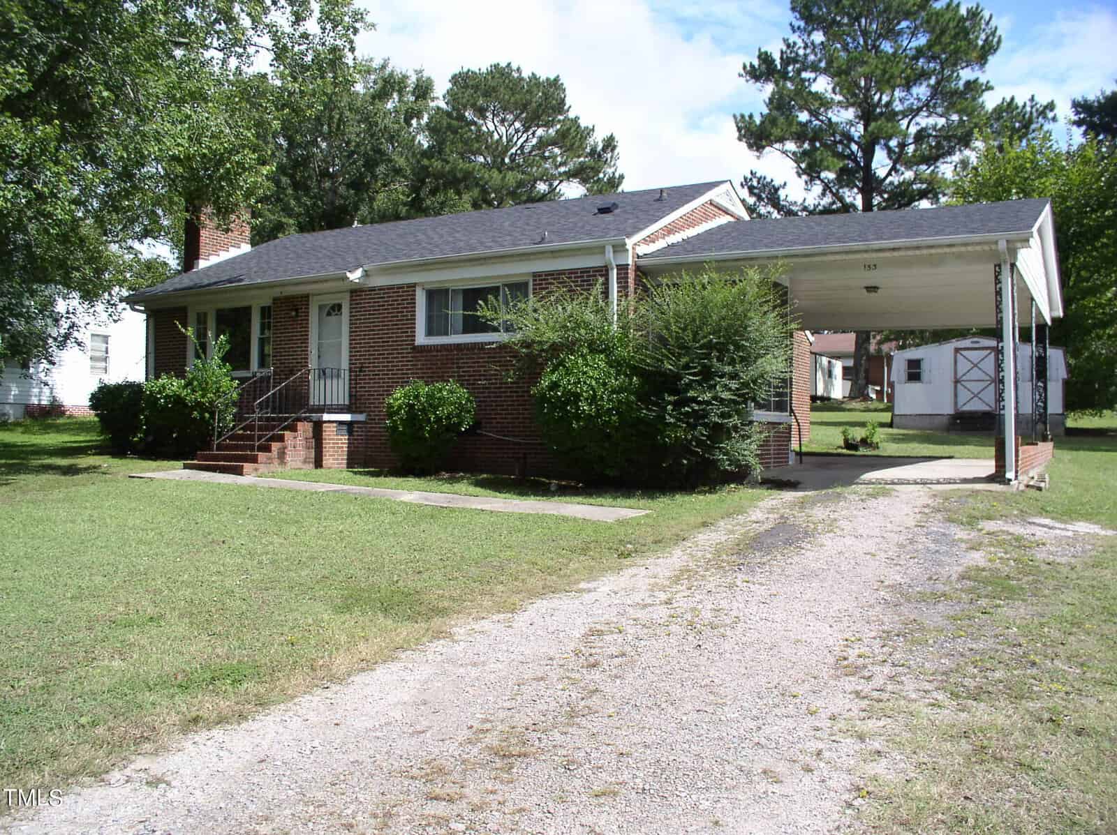 A single-story brick house at 153 S Bullock Street in Henderson features a black roof and a small porch with steps leading to the front door. A carport is attached to the right side of the house. The property is surrounded by grass and trees, with a white shed visible in the background.