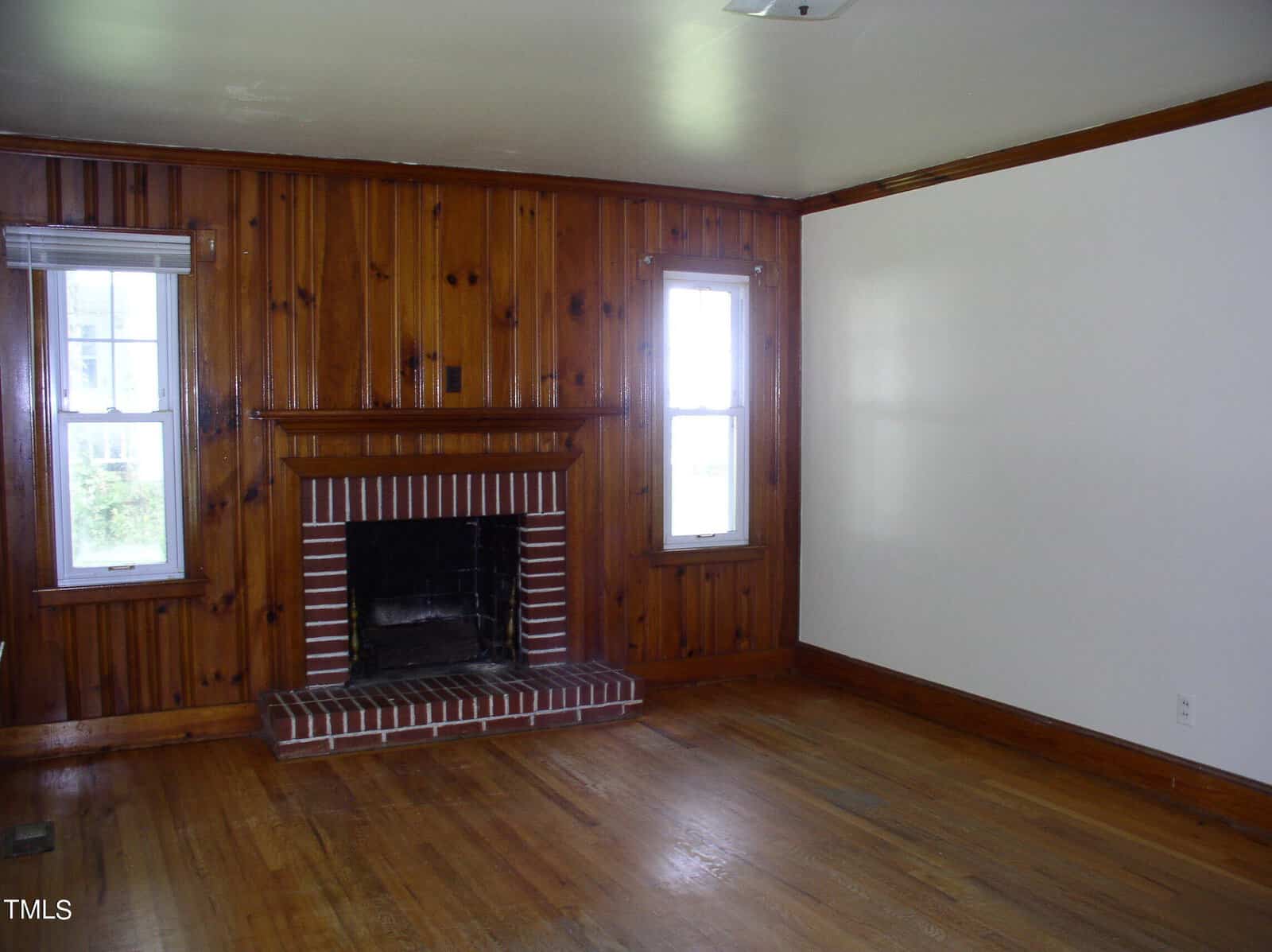 A living room with wooden floors and wood-paneled walls. A brick fireplace with a wooden mantel sits between two windows. The ceiling and one wall are painted white. The room is empty and well-lit by natural light from the windows.